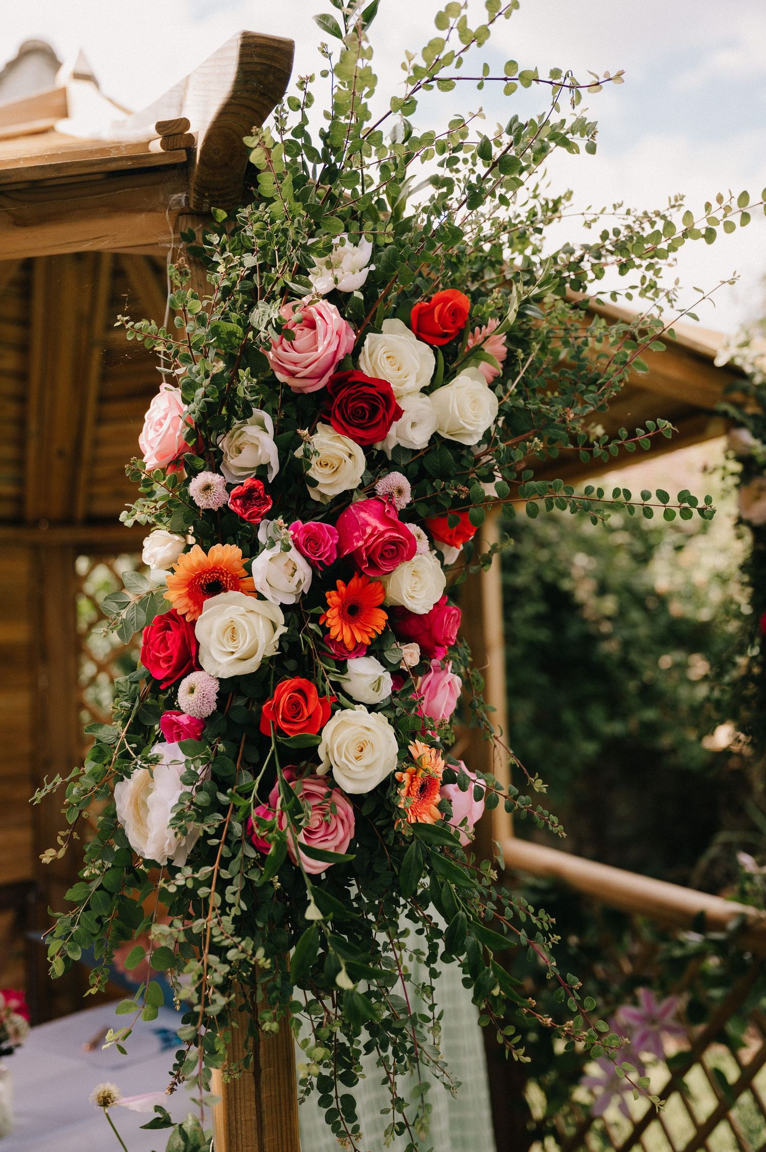 Colorful wedding arch adorned with red, pink, white roses, gerbera daisies, and lush greenery at an outdoor ceremony.