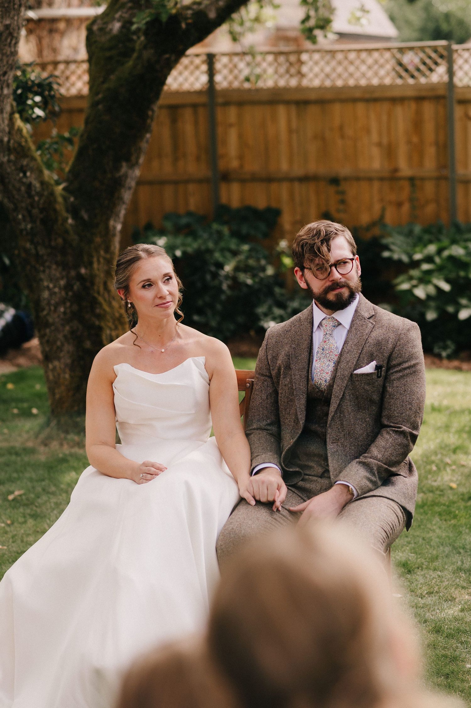 Bride in strapless white gown and groom in brown tweed suit sit together during outdoor wedding ceremony.