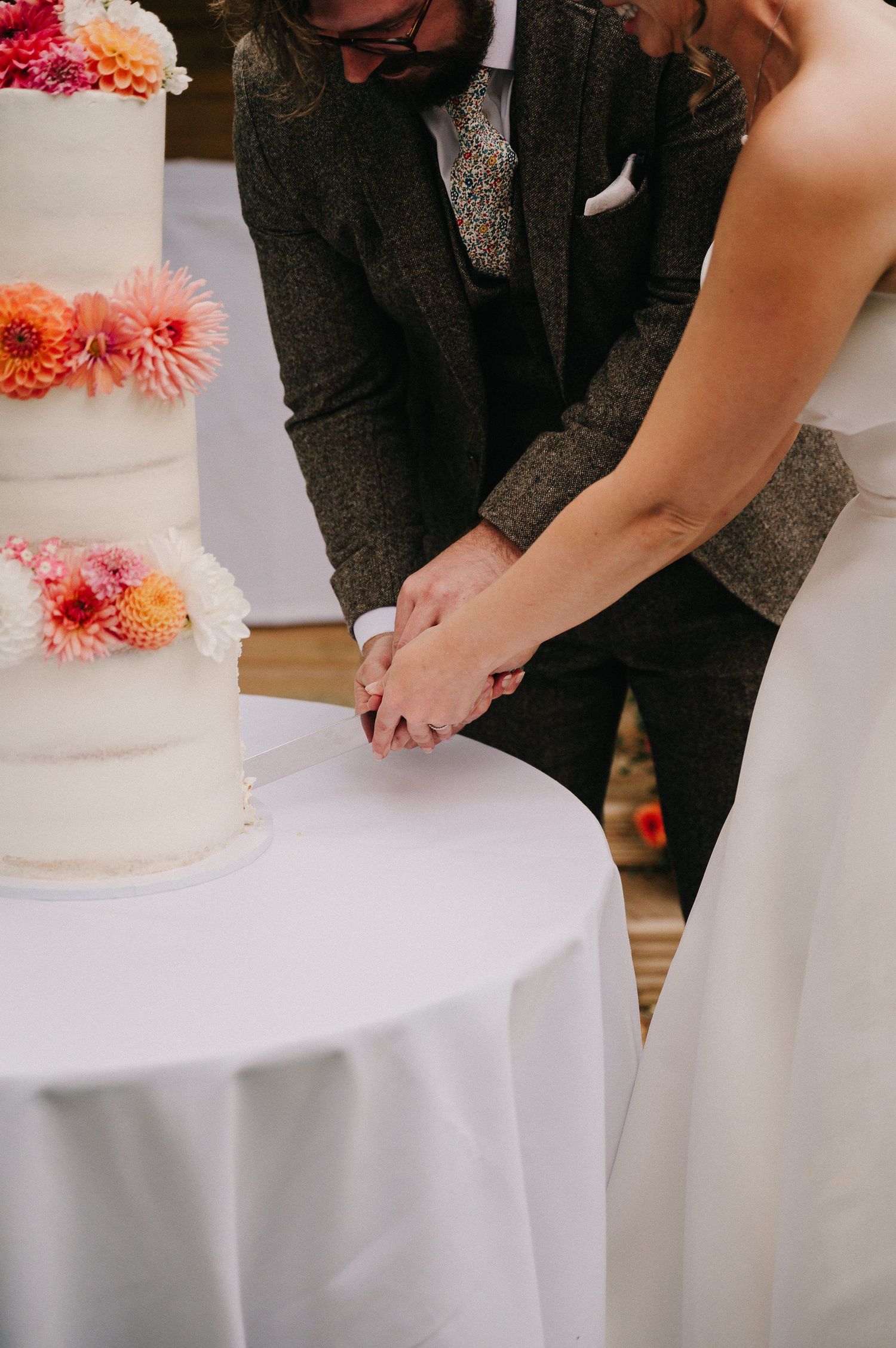 Bride and groom cutting white tiered wedding cake decorated with colorful pink and orange dahlias.