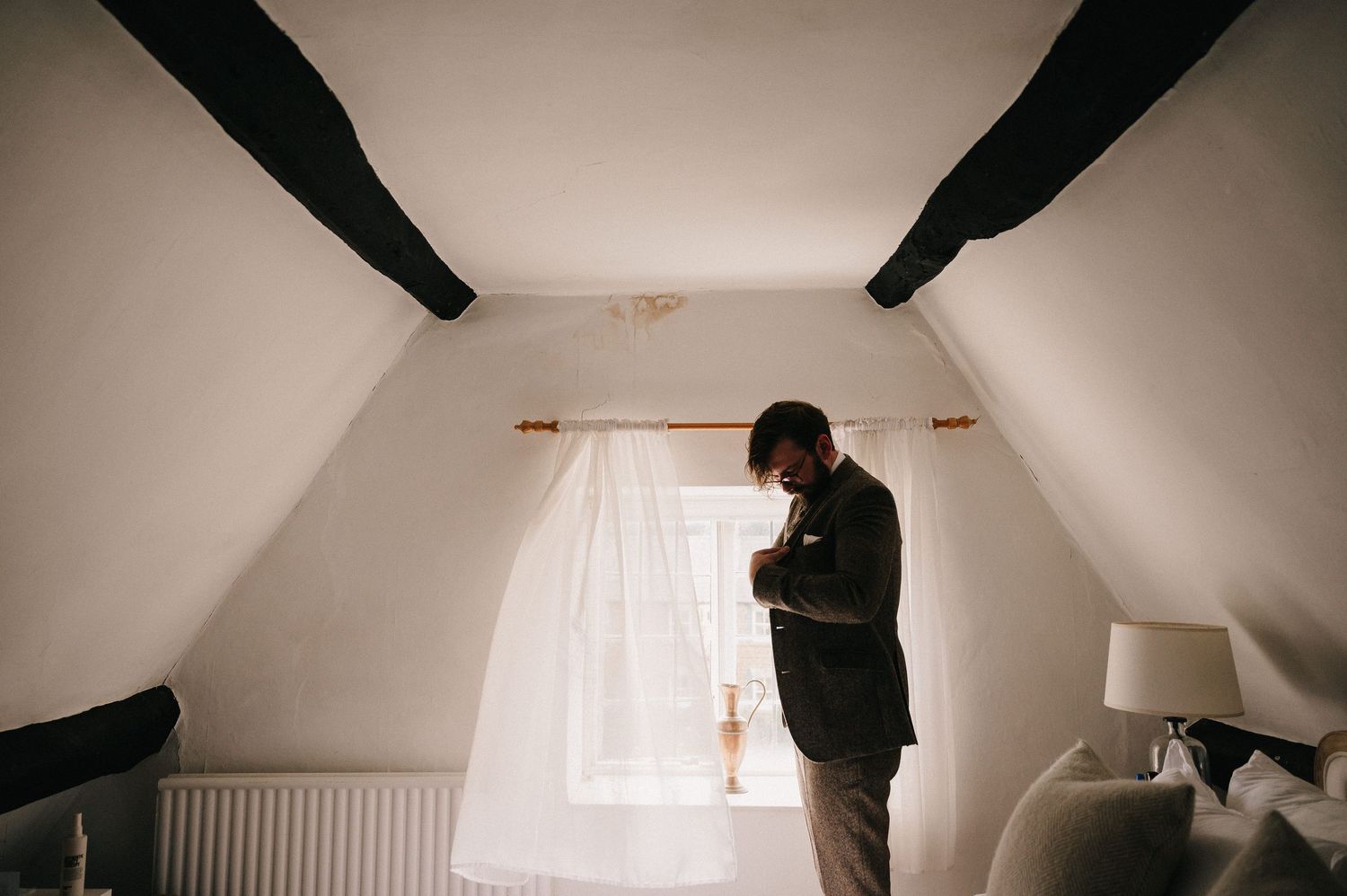 Groom standing by attic window in suit, adjusting boutonniere in soft natural light during wedding preparations.