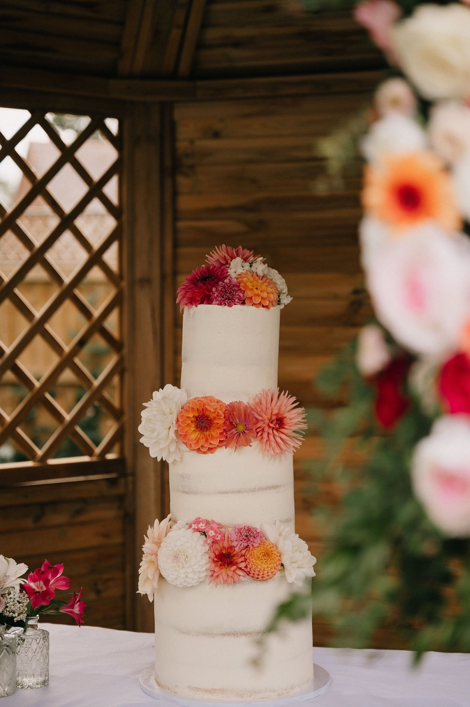 Four-tier white wedding cake adorned with colorful pink, orange, and red dahlias displayed in a wooden gazebo.