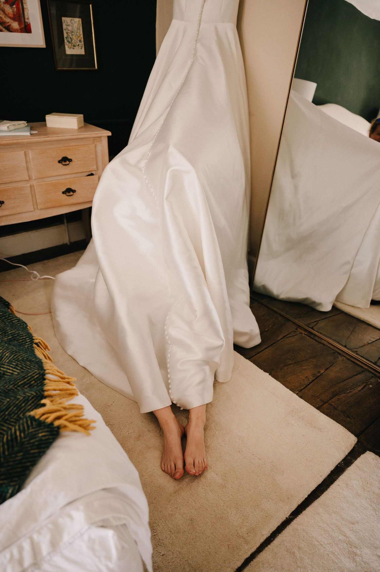 Bride in a flowing white wedding gown standing barefoot on a white rug in a bedroom with a mirror nearby.