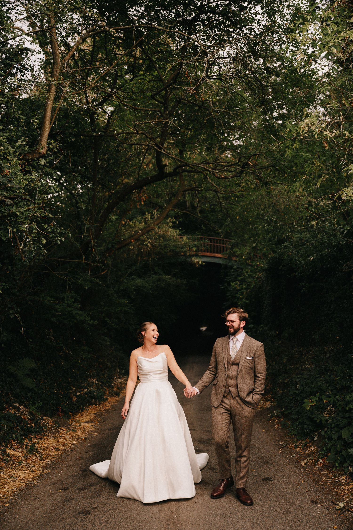 Bride in white gown and groom in brown suit hold hands on a tree-lined path in a lush garden setting.