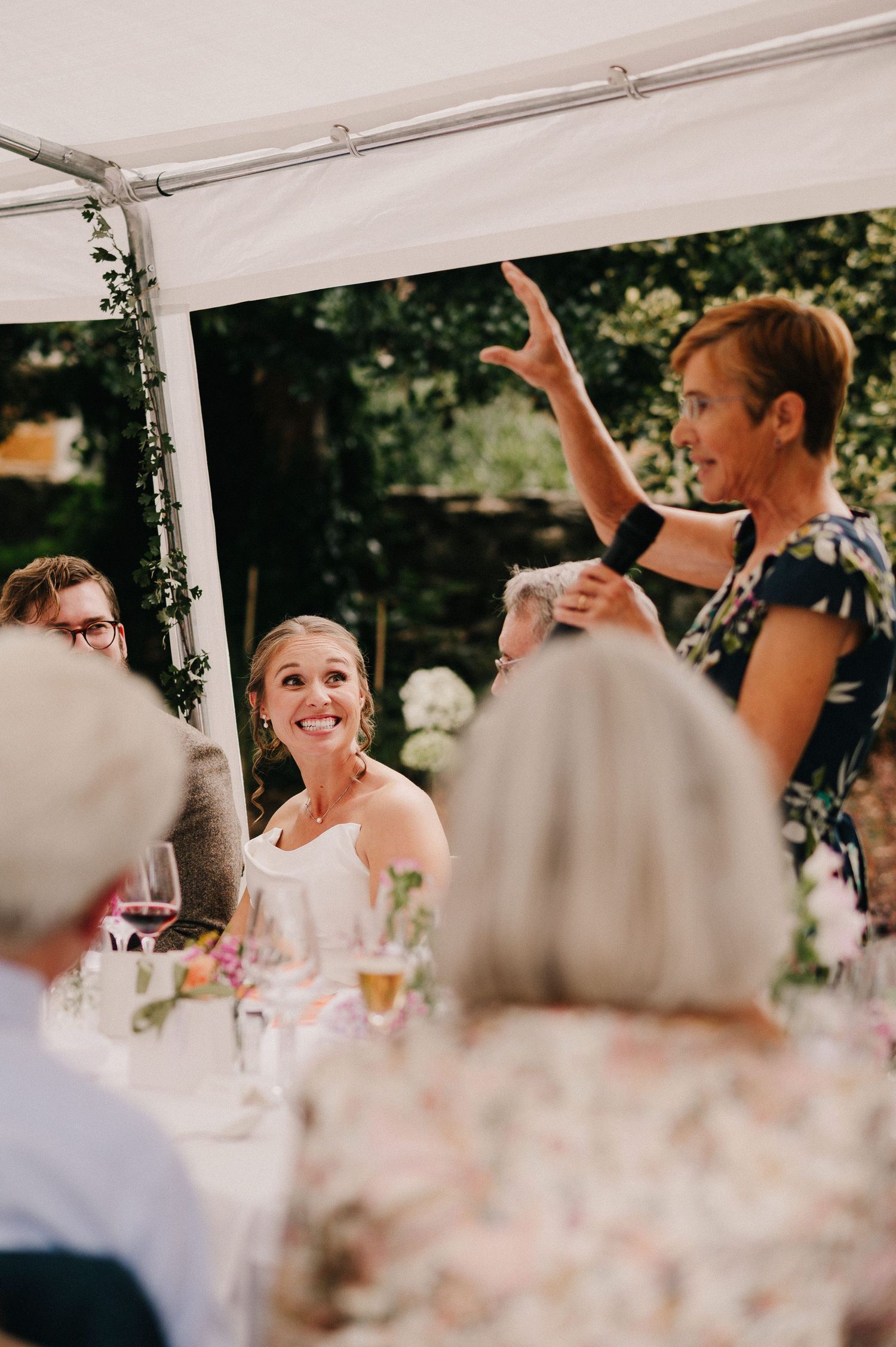 Smiling bride at outdoor wedding reception under white tent while guest gestures expressively.