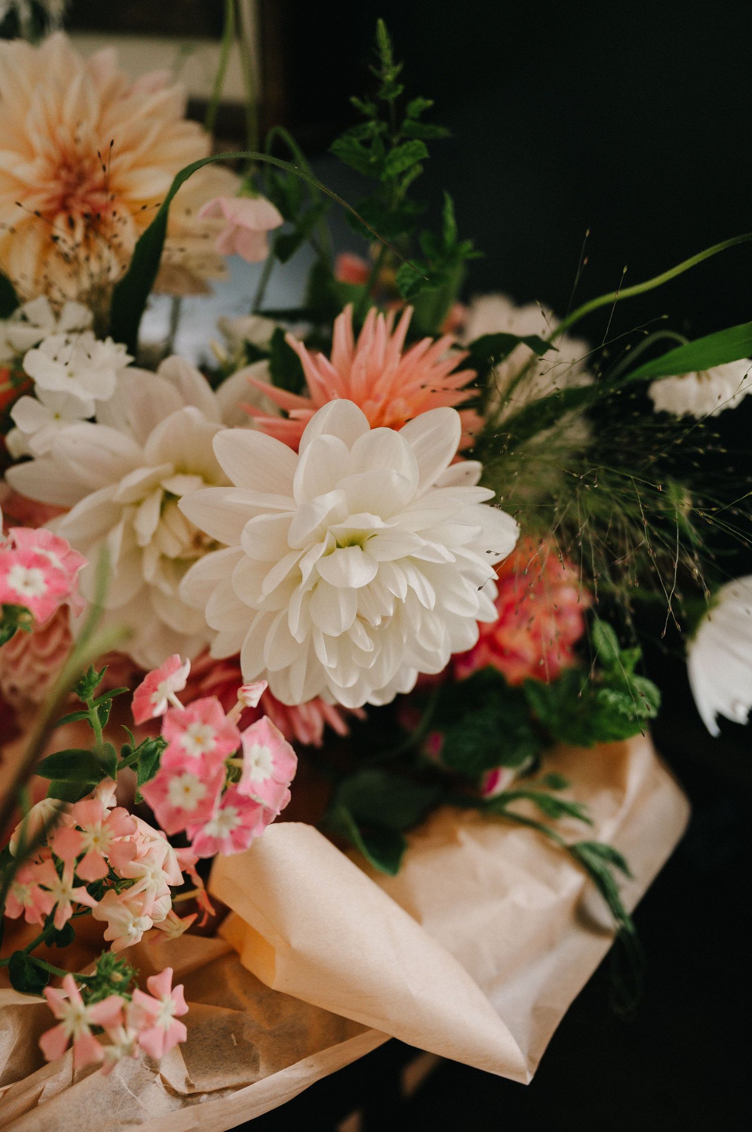 A lush bouquet of white dahlias, pink flowers, and greenery wrapped in kraft paper.