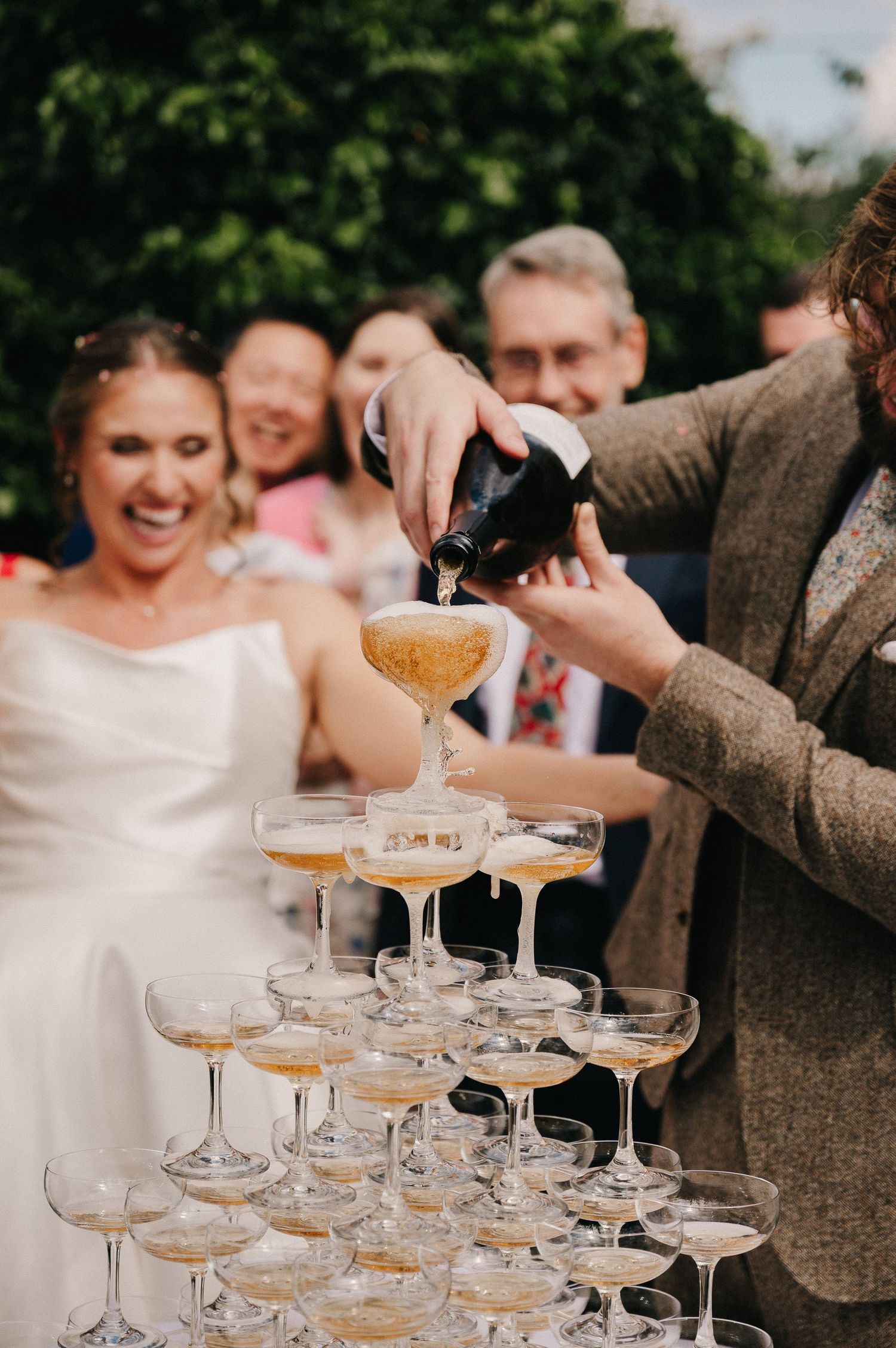 Champagne tower being poured at a wedding reception with a laughing bride in a white dress in the background.