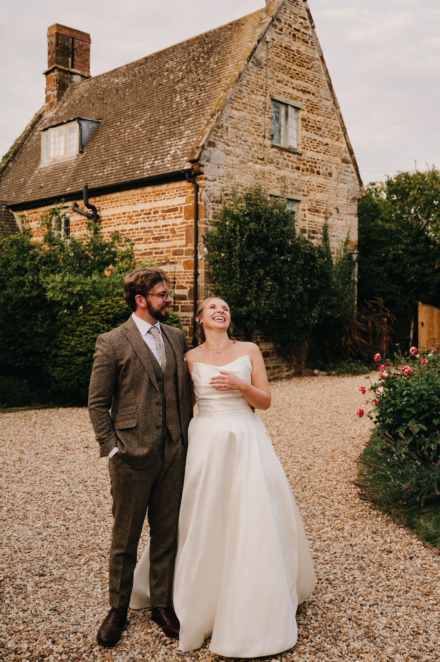 A bride and groom pose together on a gravel path outside a rustic stone farmhouse on their wedding day.