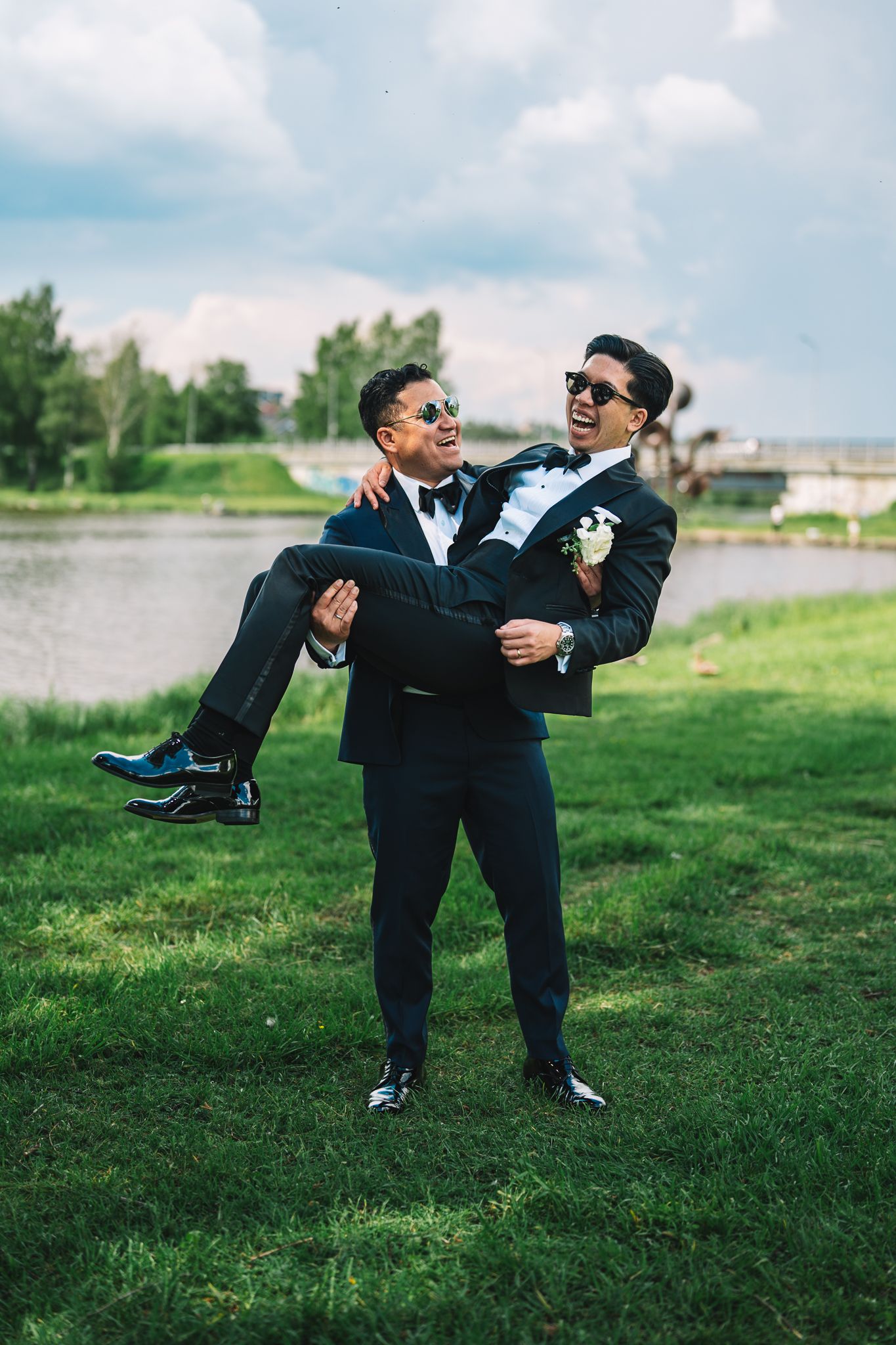 Two people in formal attire playfully dancing on a grassy lawn near a lake.