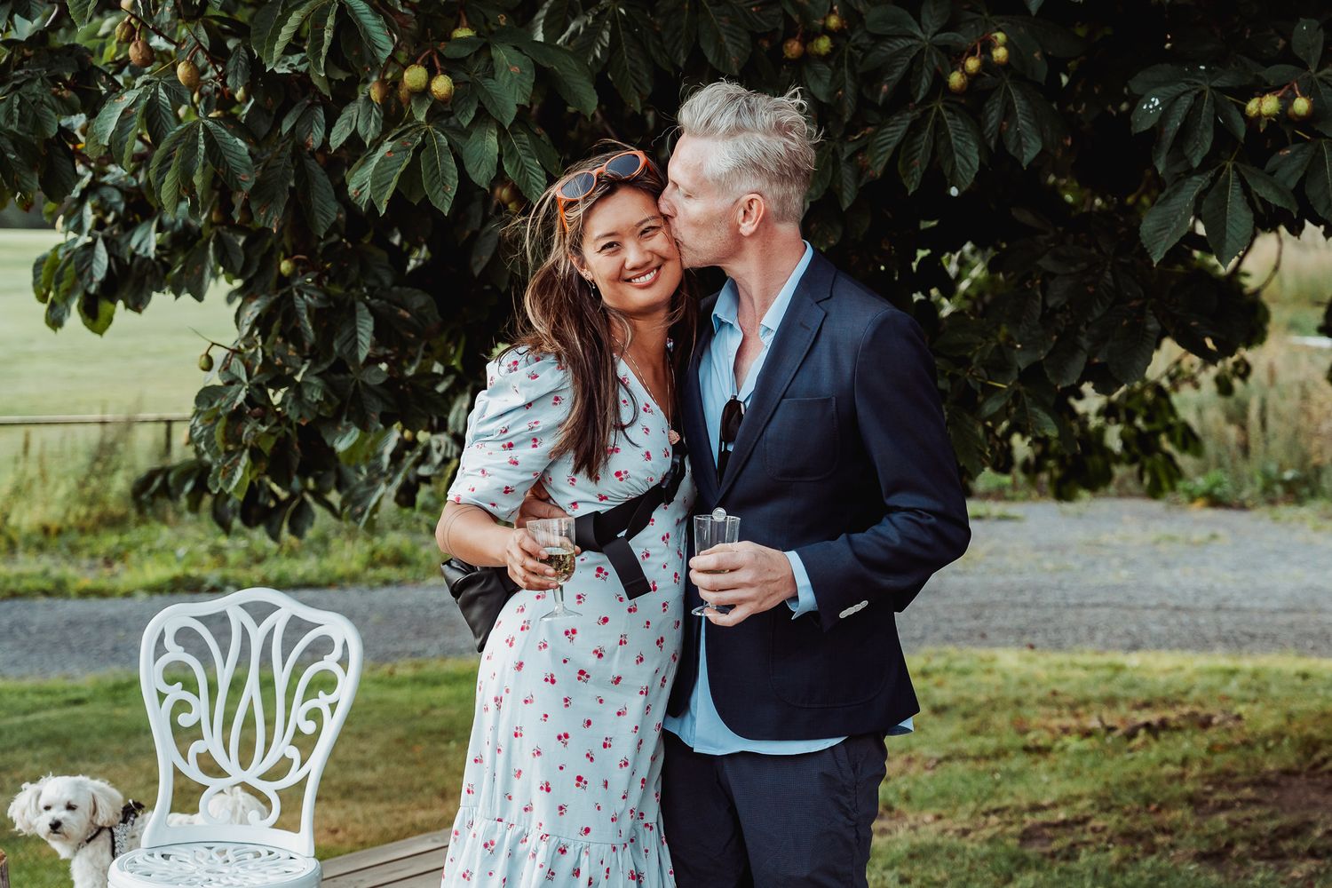 A couple shares a joyful moment outdoors near a decorative white chair, with the woman wearing a polka dot dress.