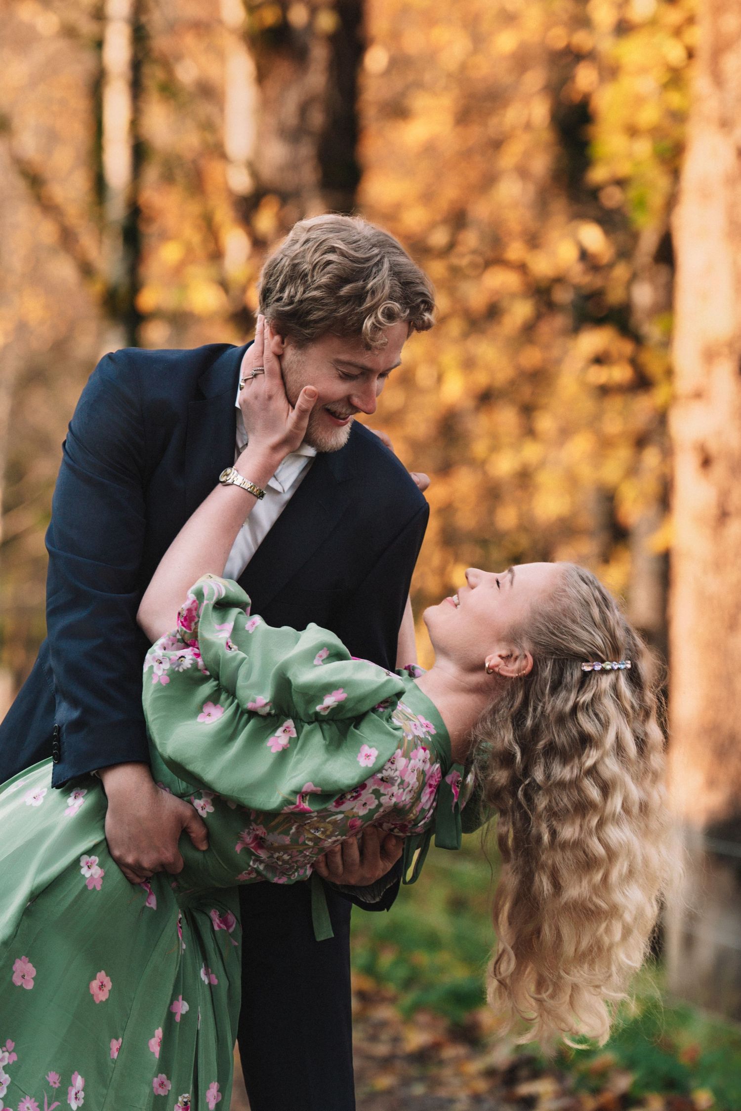 Romantic autumn engagement photo with couple in green dress and suit among fall foliage.