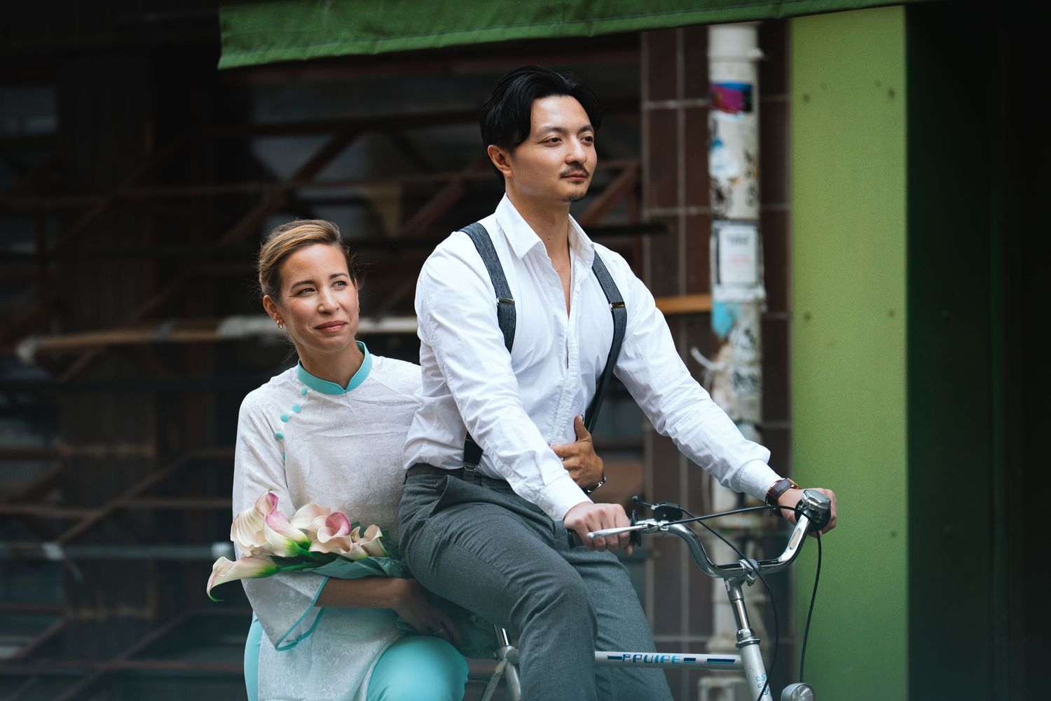 Two people ride together on a bicycle along a city street with green buildings in the background.