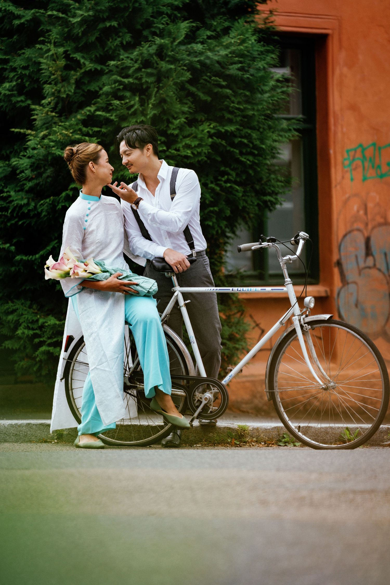 Romantic couple in light colored clothing share tender moments near a vintage bicycle against an orange wall.