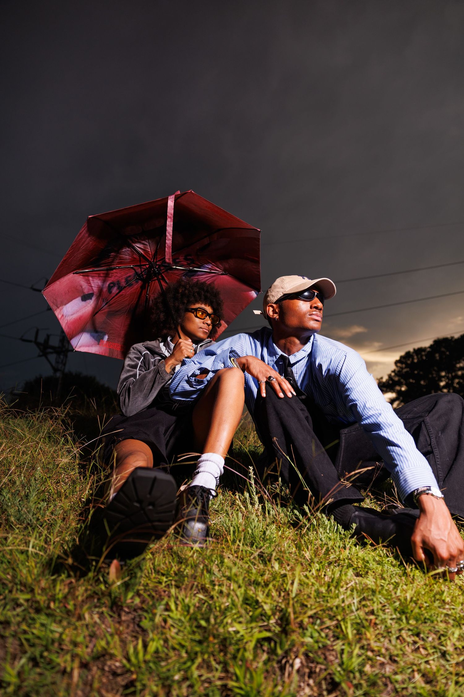 Two people sitting on grass under a red umbrella with a dramatic stormy sky backdrop.