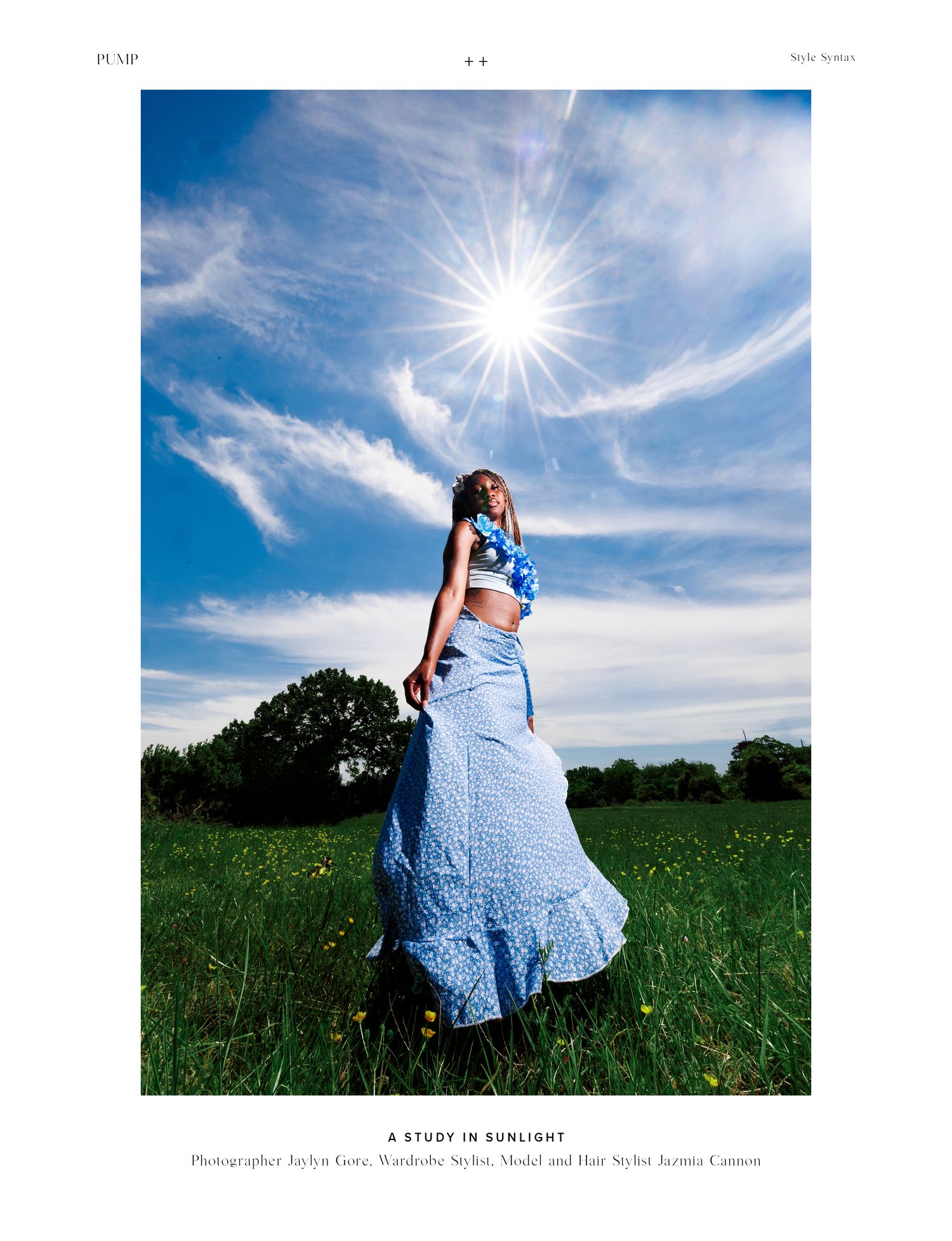 Woman in blue floral skirt and crop top poses in sunlit green field under dramatic blue sky with wispy clouds.