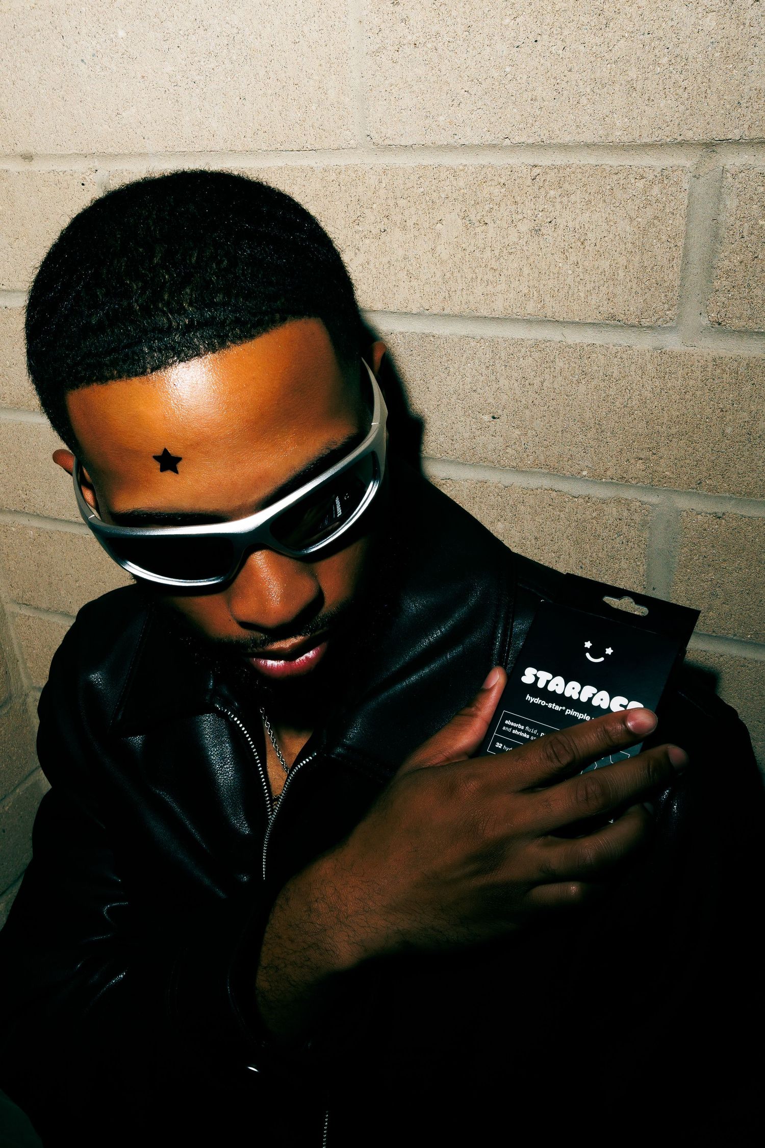 Young man in futuristic sunglasses and leather jacket holds Starface skincare product with star patch on forehead.