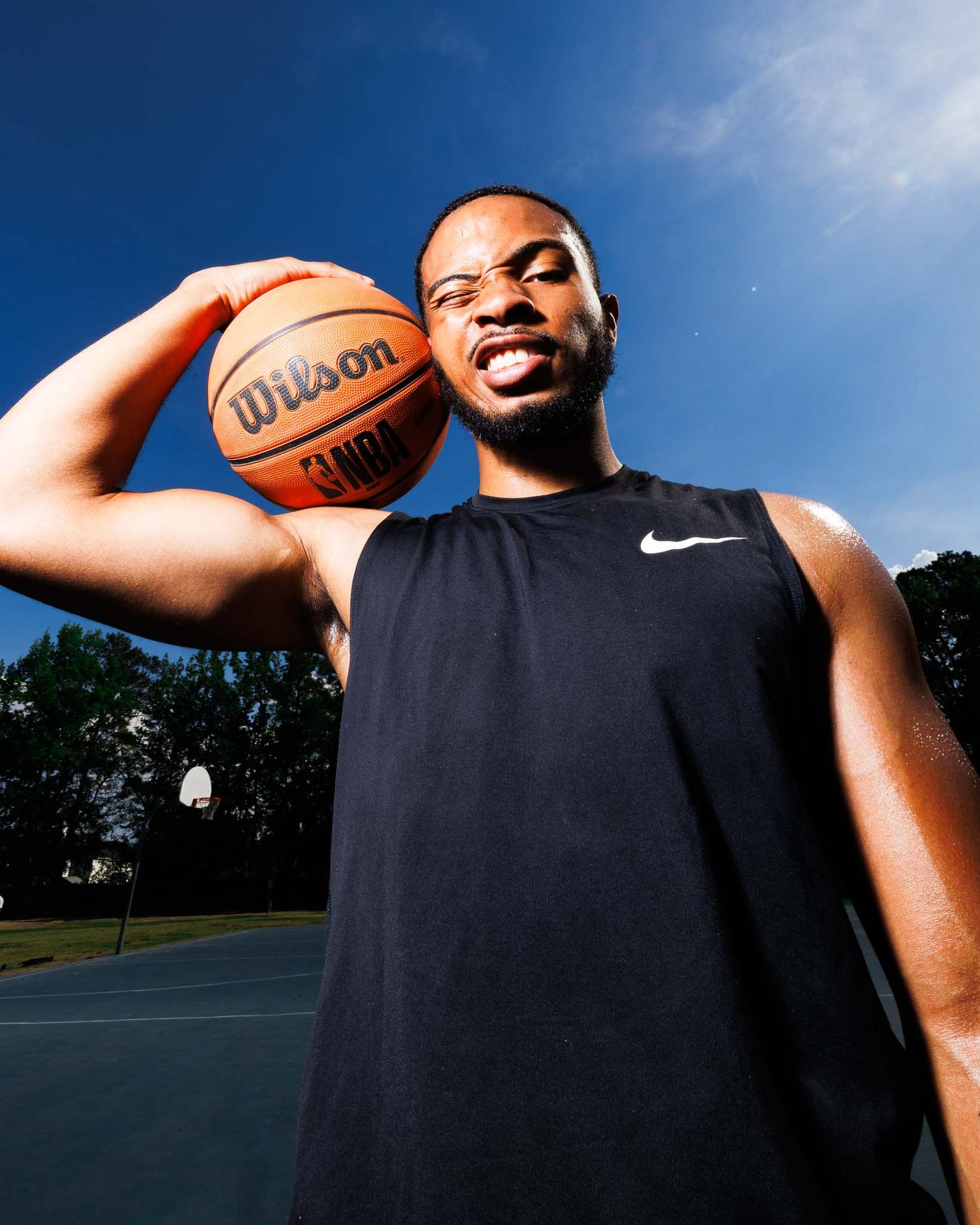 A man in a black Nike tank top holds a Wilson basketball on an outdoor court under a blue sky.