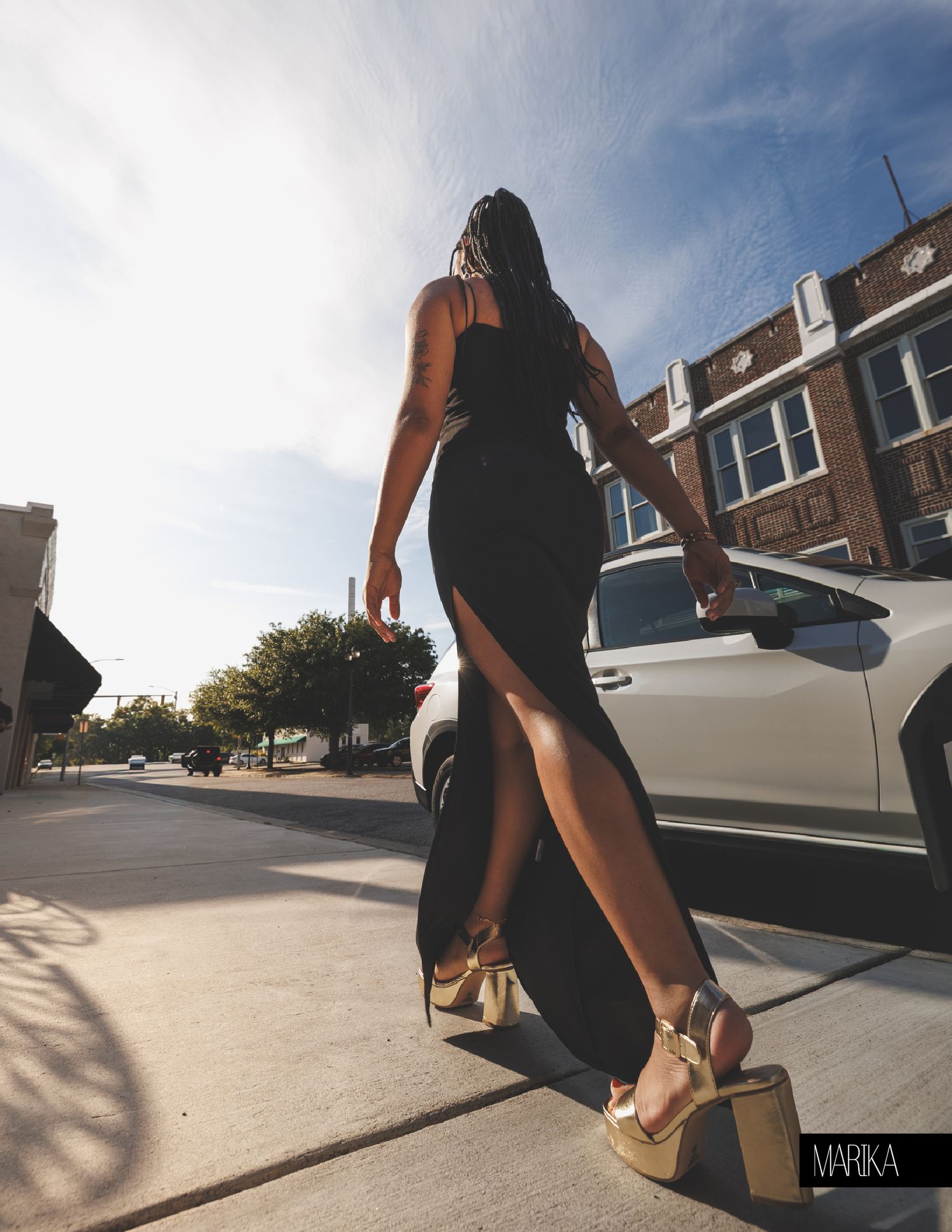 Woman in black slit maxi dress and gold platform heels walking on urban sidewalk near white car on sunny day.
