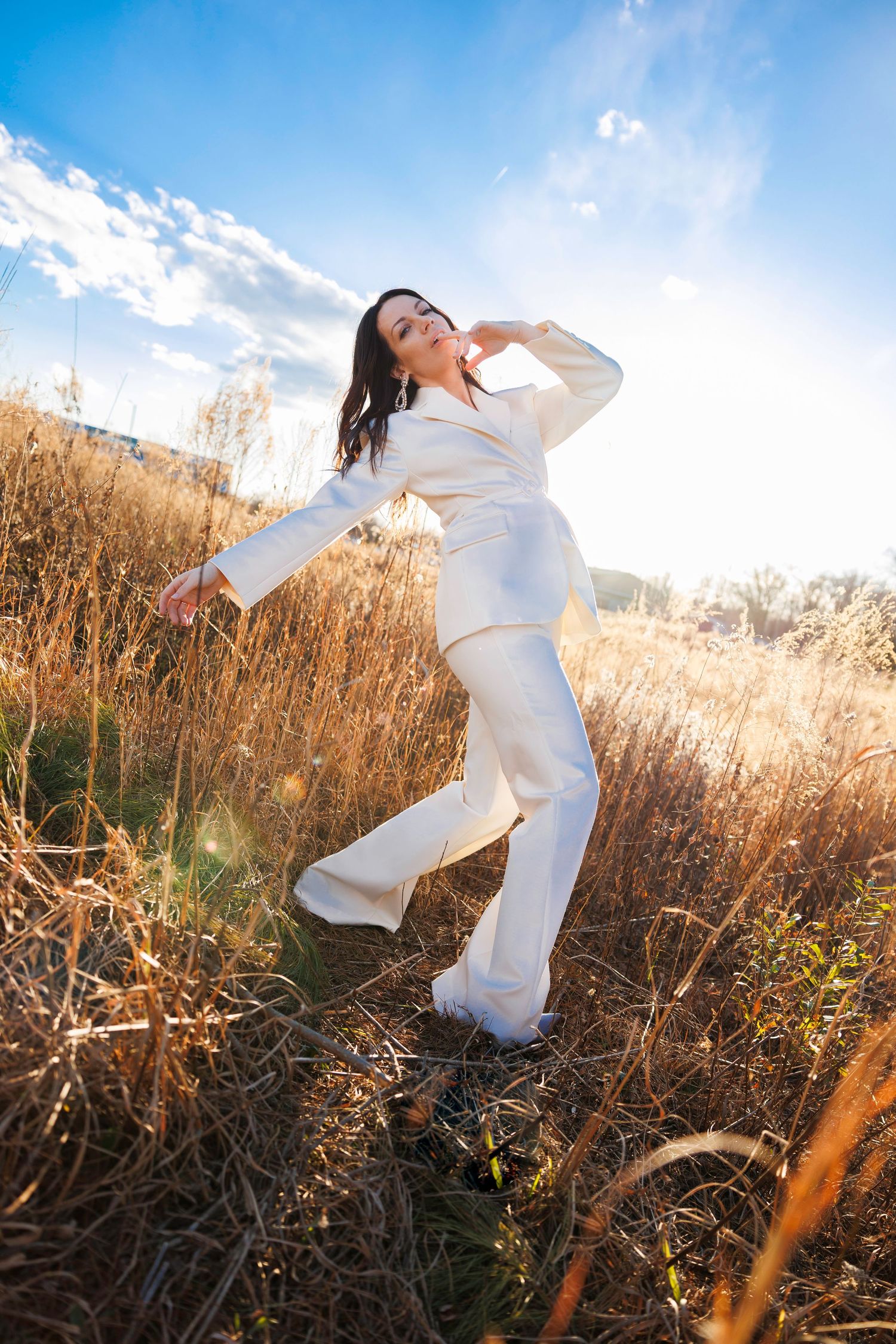 A person in a white outfit poses expressively in a field of tall golden grass against a bright blue sky.