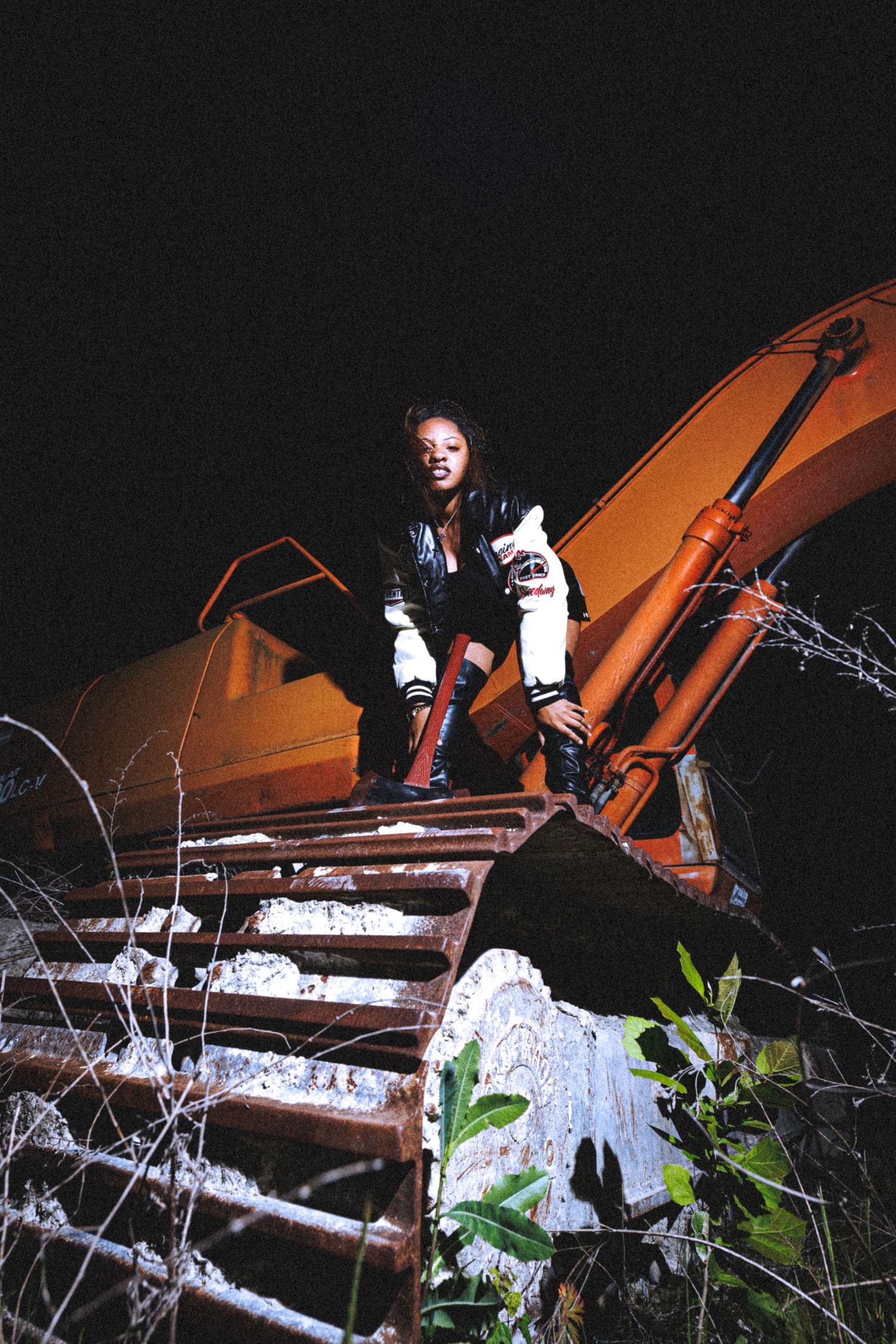 A young woman in a varsity jacket poses on rusty stairs beside orange construction equipment at night.
