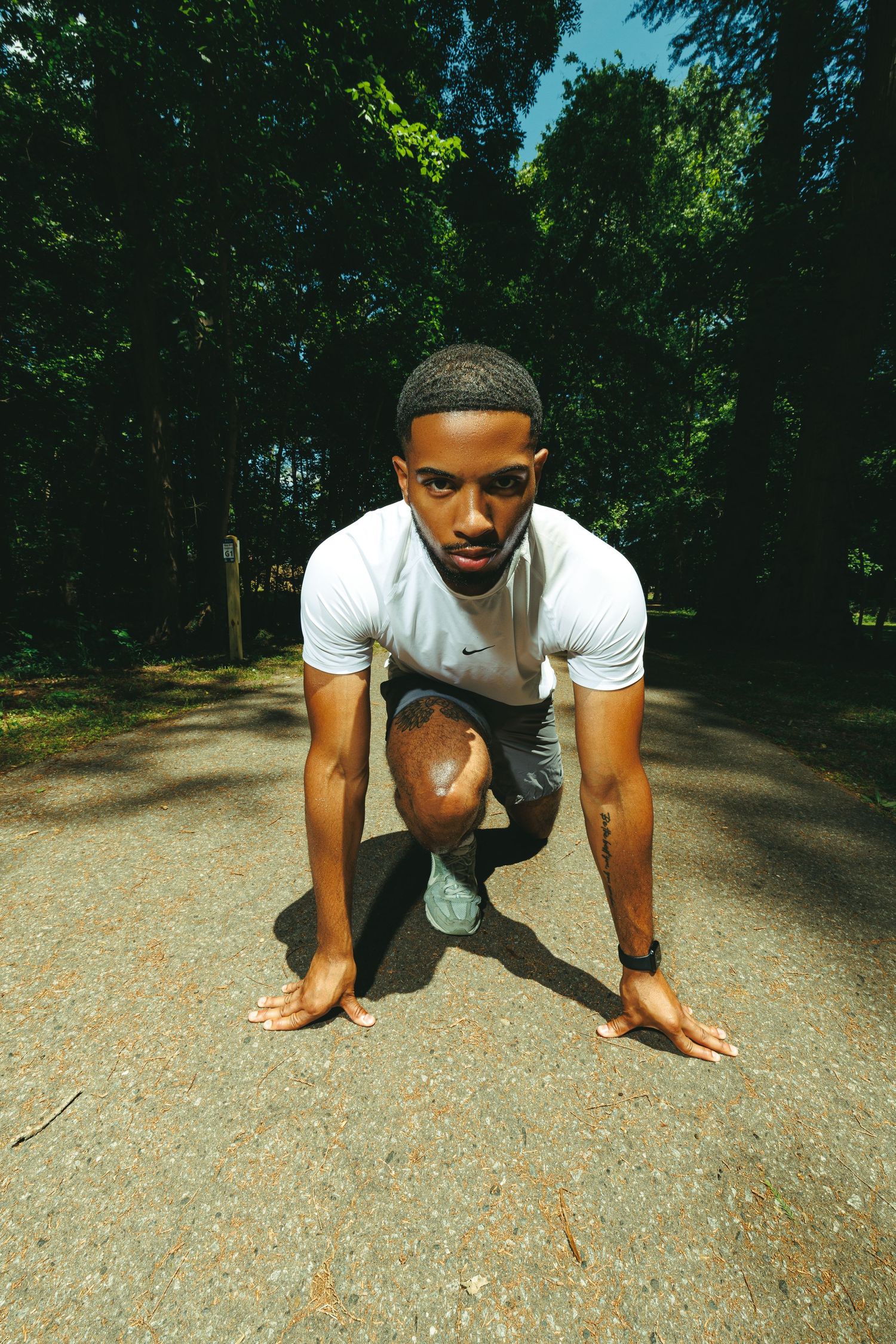 Young Black male athlete in starting position on a park path, wearing white Nike shirt and gray shorts.