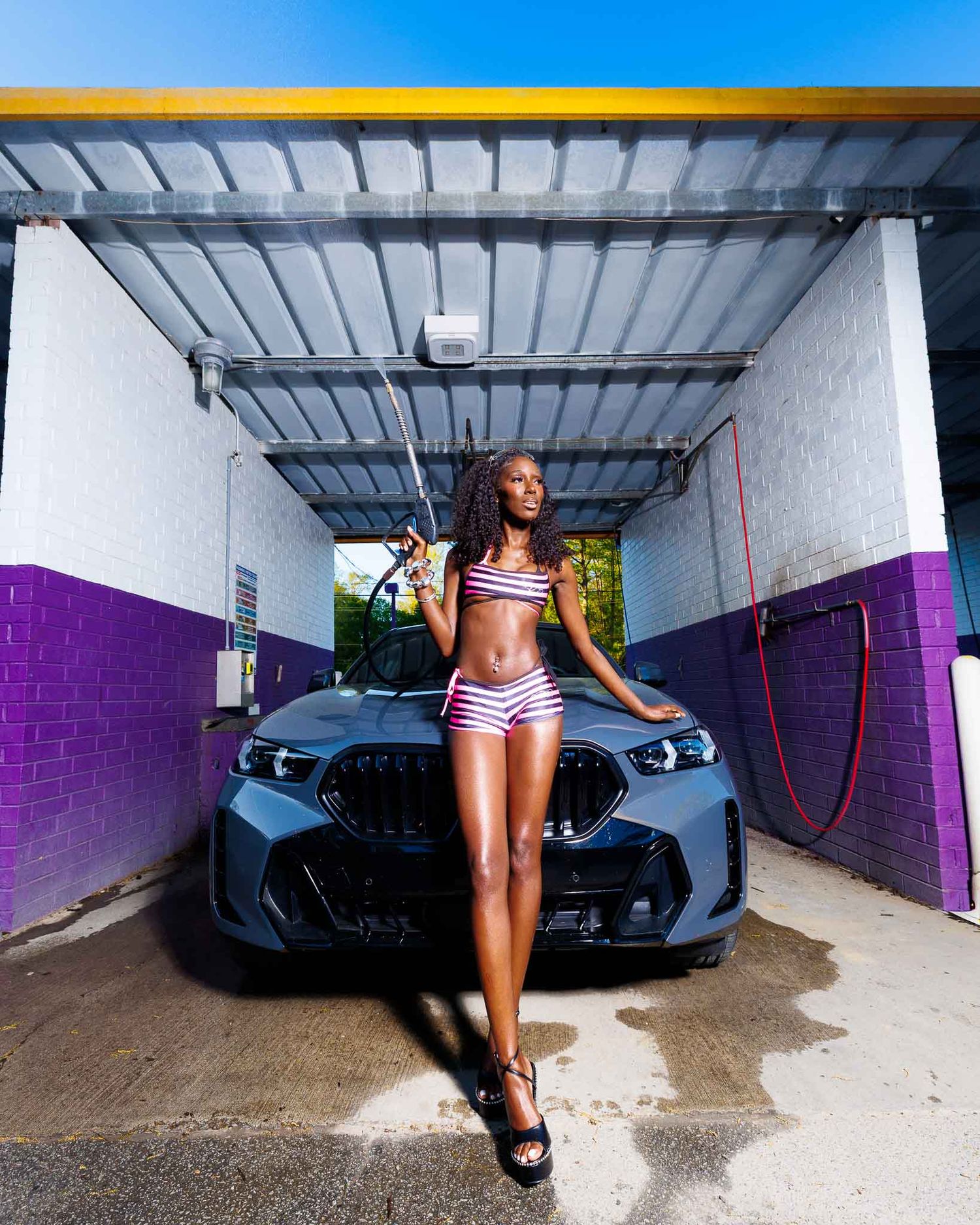 Woman in striped bikini posing on BMW car hood at purple-accented car wash bay on sunny day.