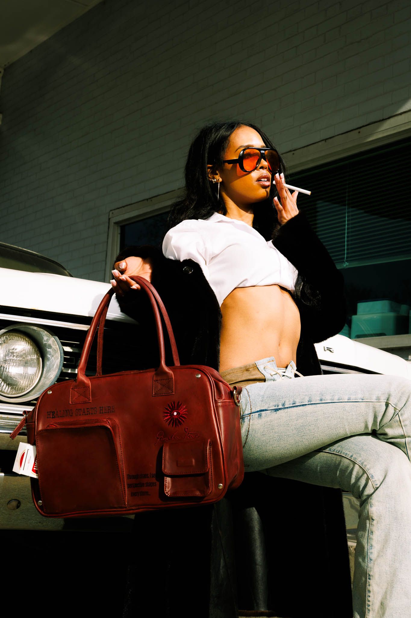 Stylish woman in white crop top and jeans posing with brown leather bag near vintage car.
