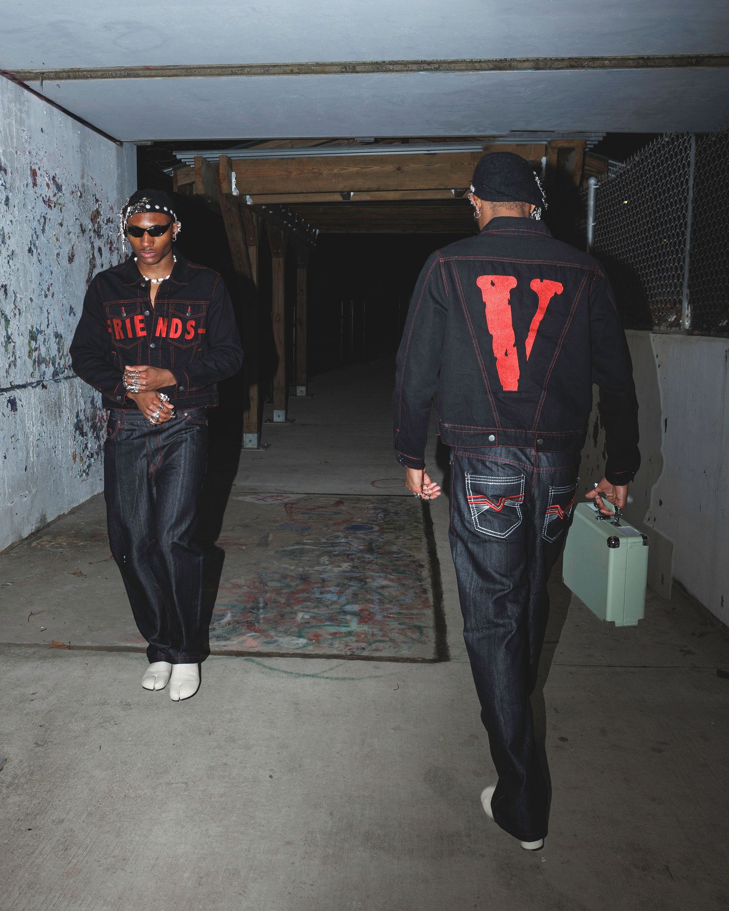 Two men in Vlone streetwear walk through an urban corridor, one wearing a black V logo jacket carrying a mint case.