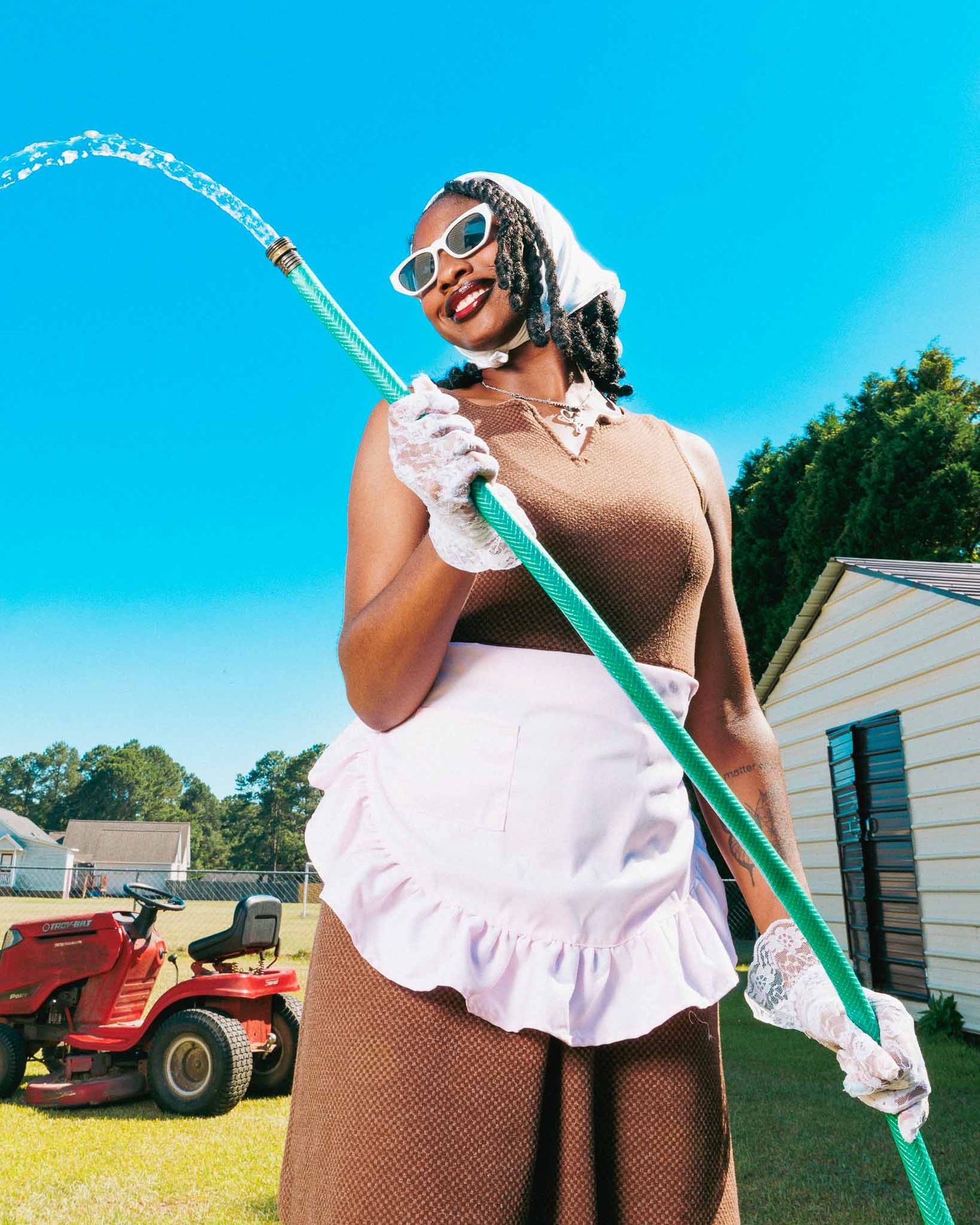 Woman in maid apron and sunglasses holds garden hose outdoors on sunny day near red lawn mower.