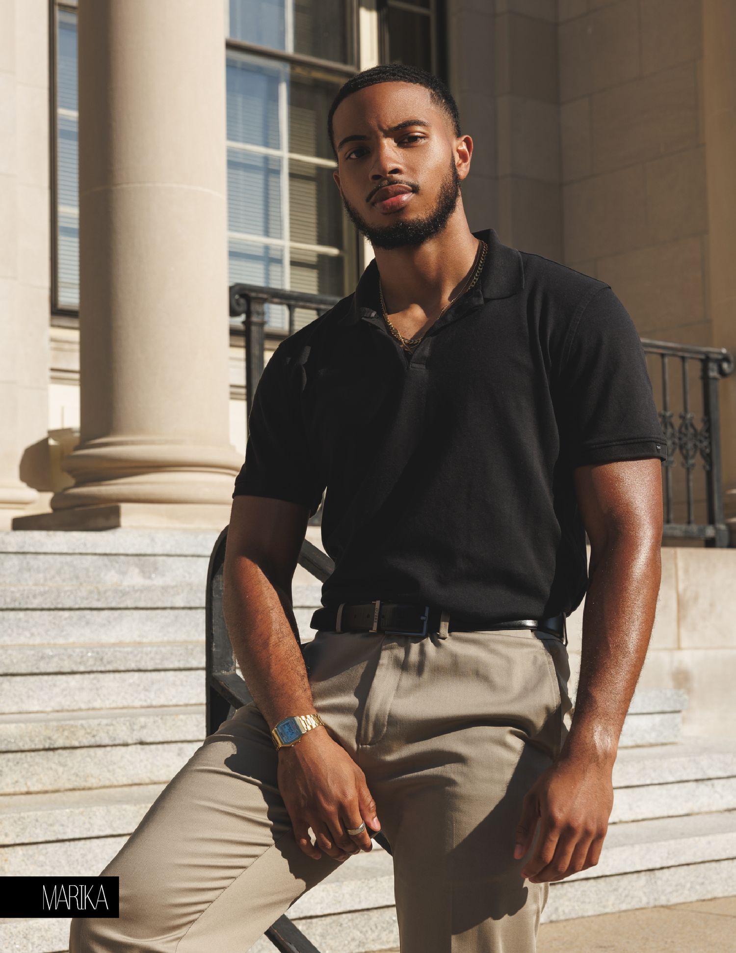 Young Black man in black polo shirt and khaki pants poses on stone steps near classical columns.