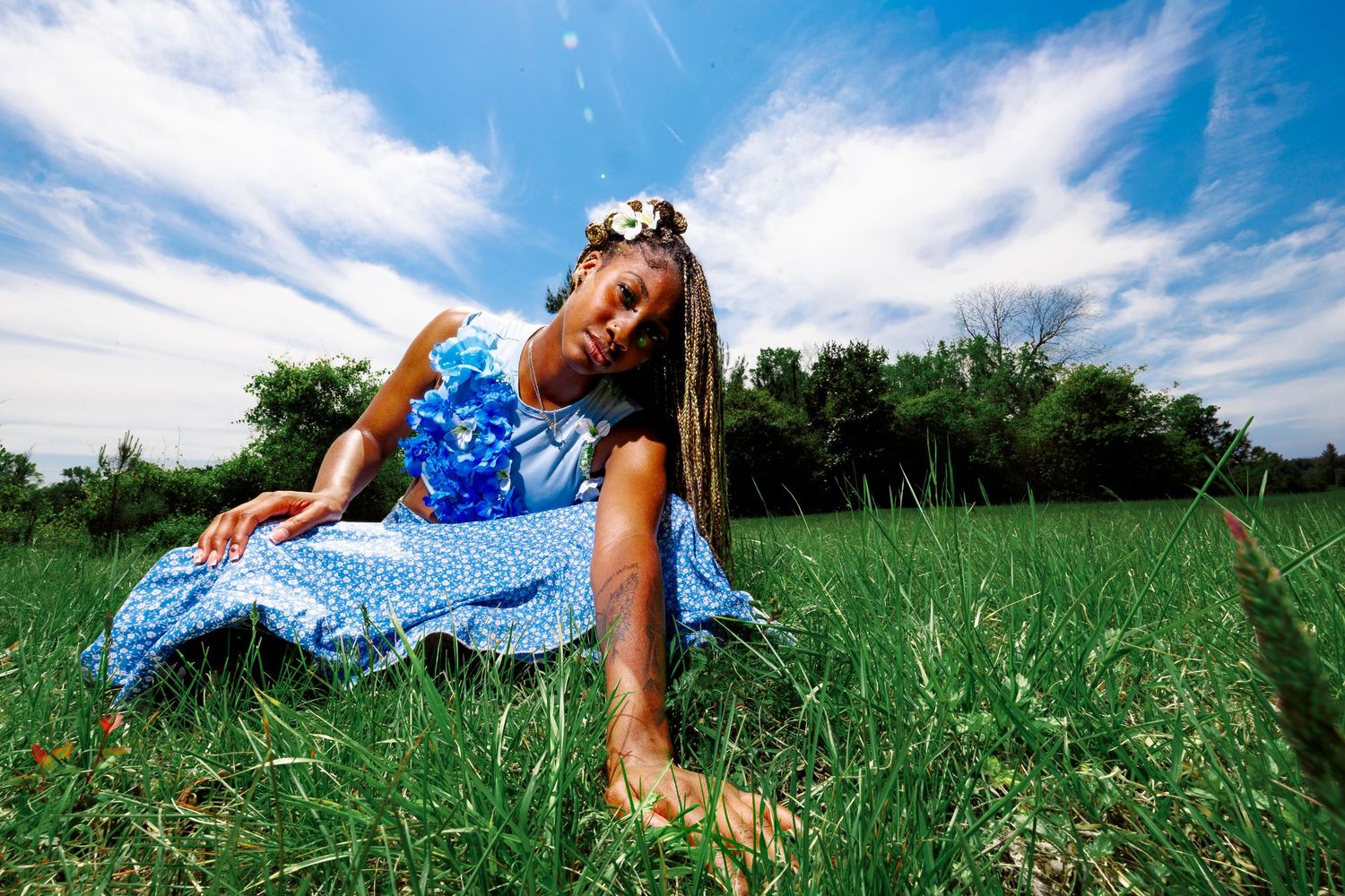 Young Black girl in blue dress and flower crown sits in lush green field under bright blue sky.