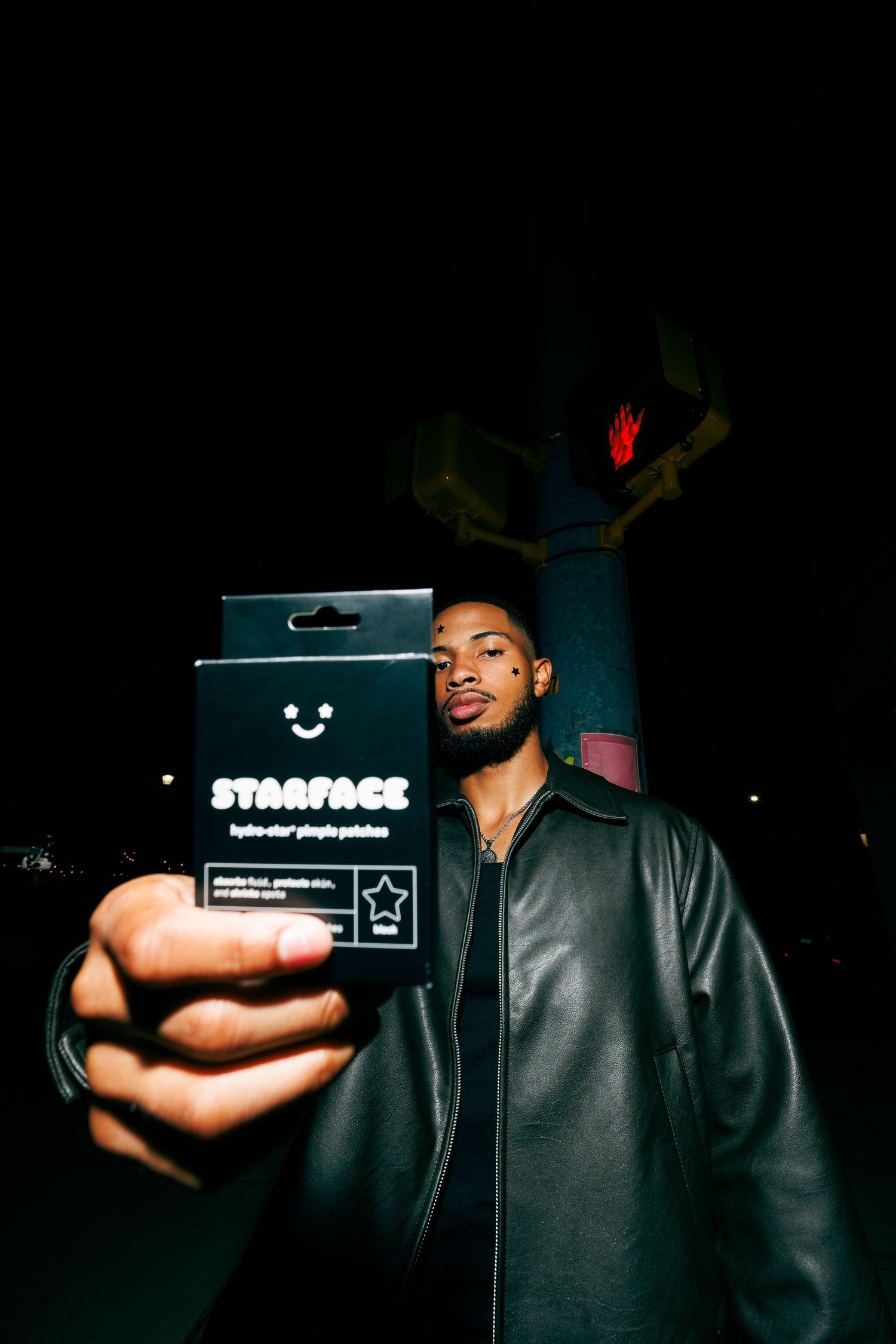 Man in black leather jacket holding Starface pimple patches box at night outdoors.
