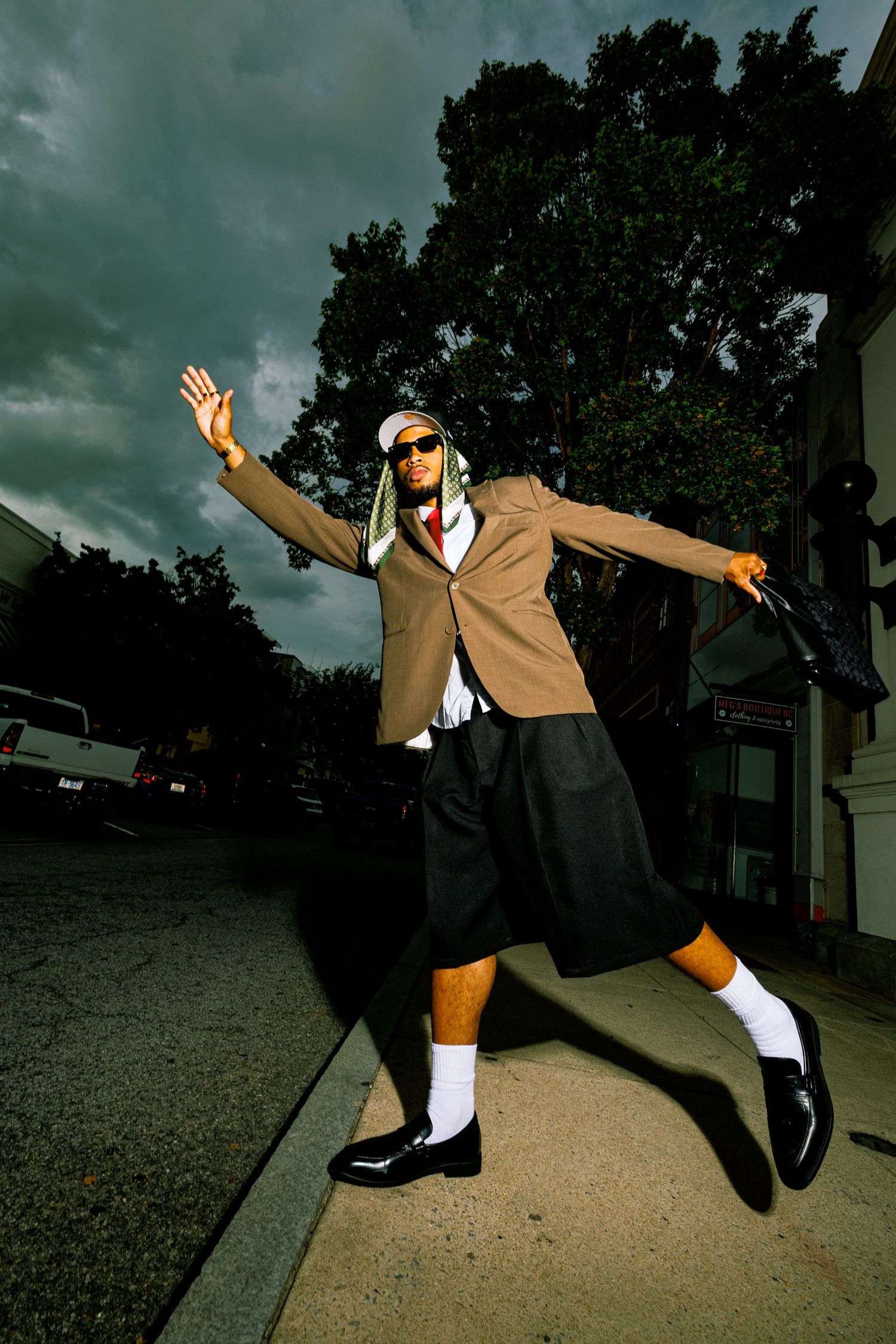 Stylish man in blazer, shorts, white socks, and black shoes poses dramatically on sidewalk under stormy sky.