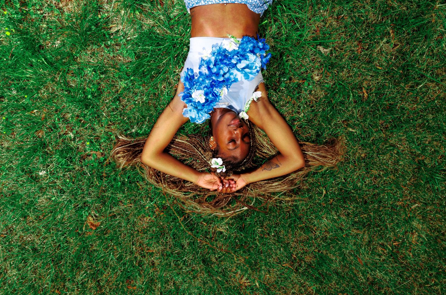 A young girl in a blue floral outfit lies on lush green grass, laughing joyfully with her hair spread around her.