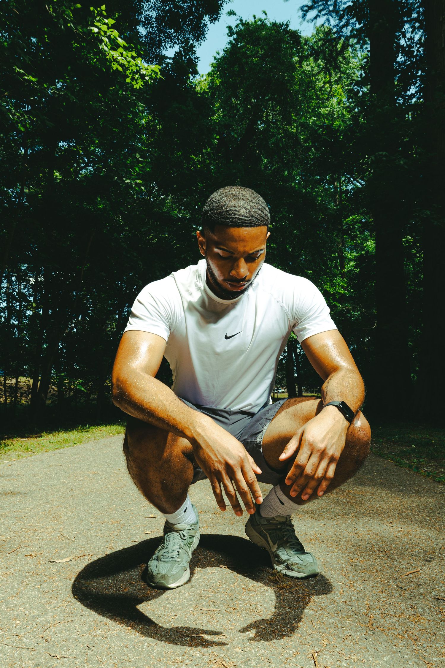 Athletic man in white Nike shirt crouching on a path in a sunlit park, wearing gray shorts and sneakers.