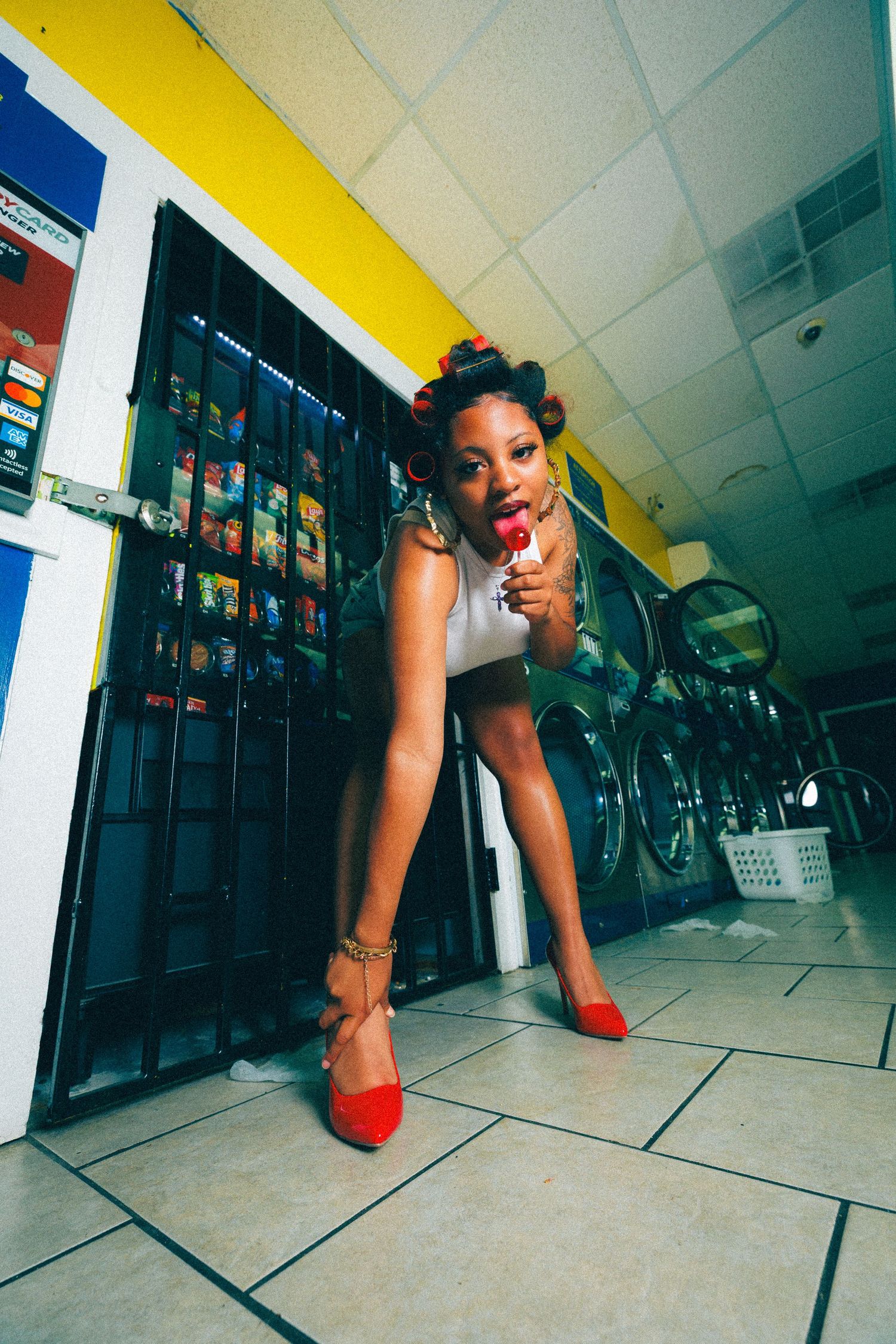 Woman in red heels and hair rollers posing in a laundromat while holding a lollipop.
