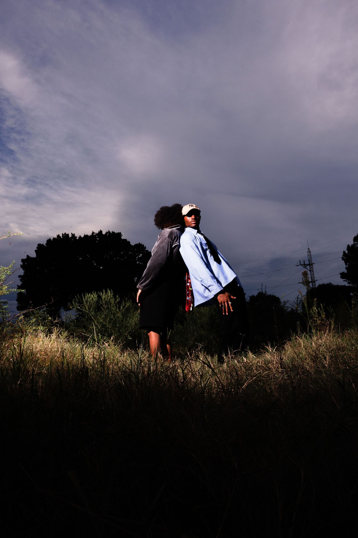 A person stands in a dark field under dramatic cloudy skies, wearing a light blue jacket and white cap.
