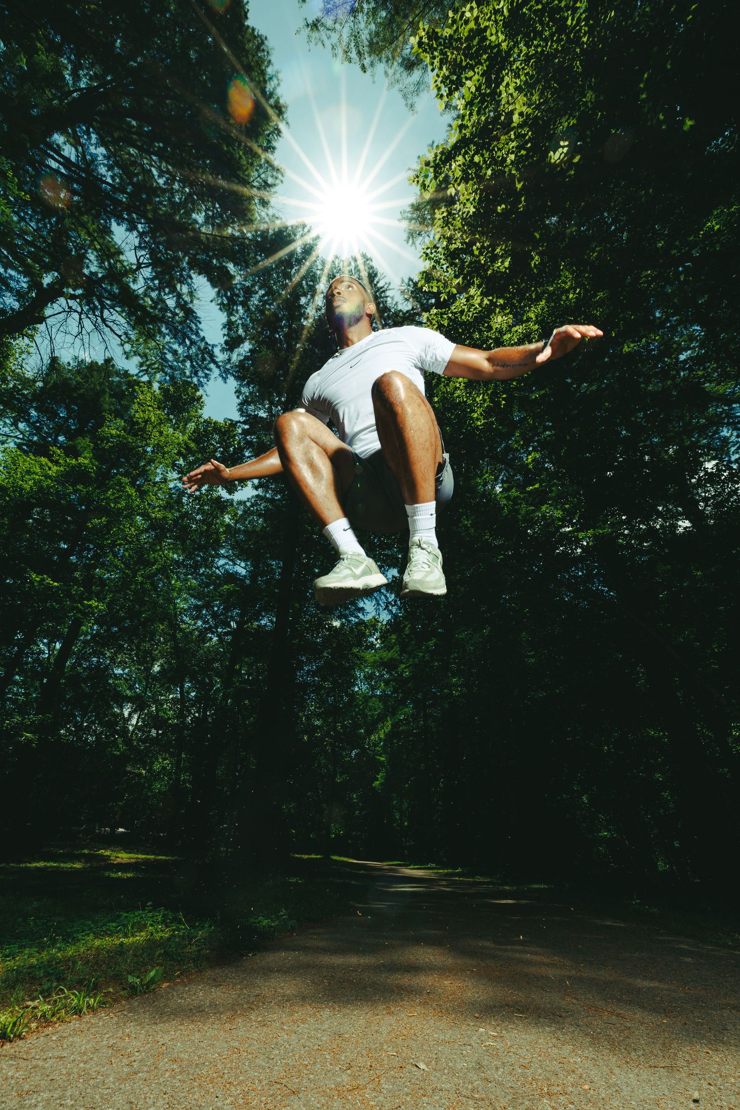 Person jumping mid-air on a sunlit park path surrounded by lush green trees with sun flare overhead.
