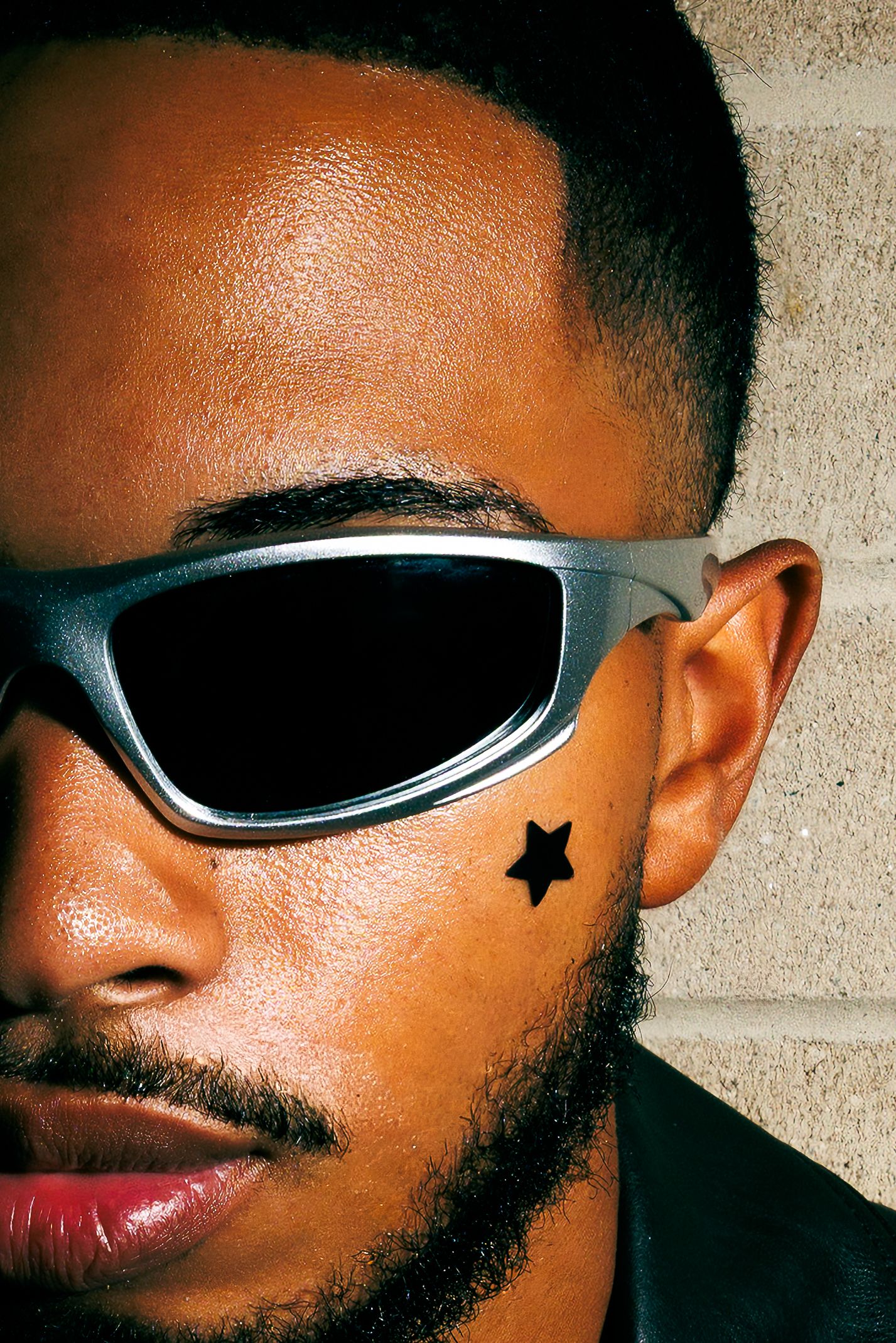 Close-up of a man with a black star face tattoo wearing silver wraparound sunglasses against a stone wall.