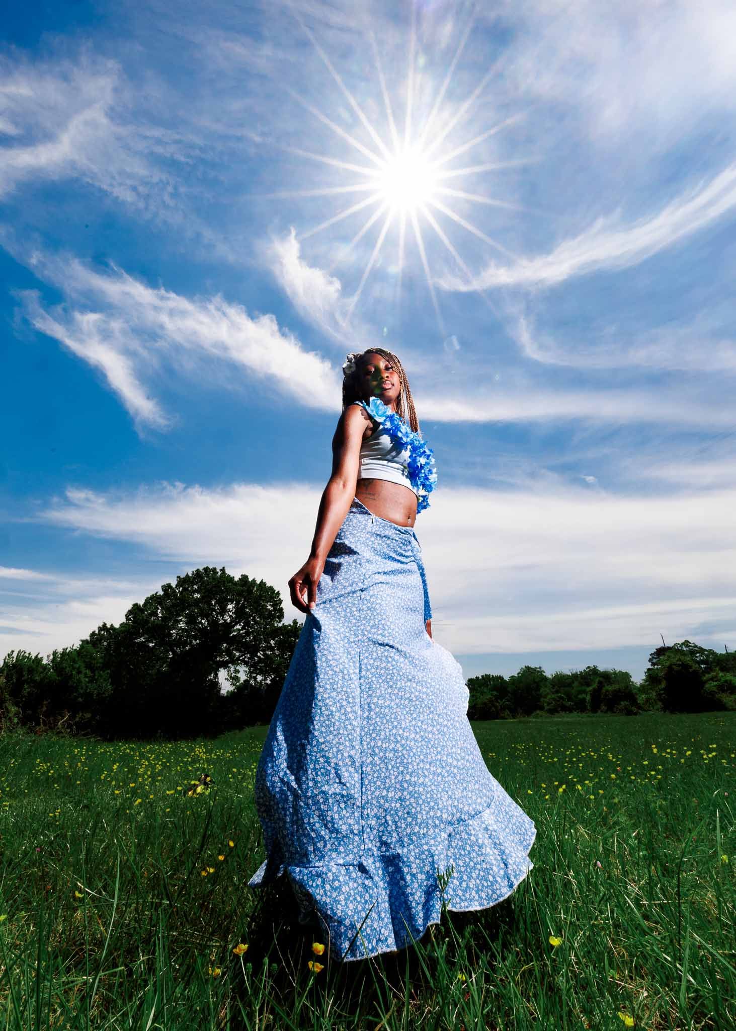 Woman in blue floral maxi skirt and crop top stands in sunny green field under bright blue sky with wispy clouds.