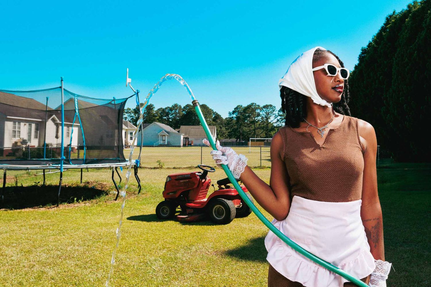 Woman in sunhat and sunglasses holding garden hose in sunny backyard with trampoline and red lawn mower.