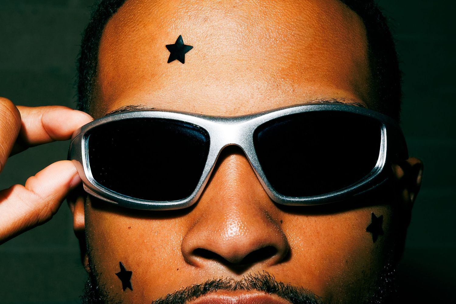Close-up of a man wearing silver sunglasses with star tattoos on his forehead and cheek.