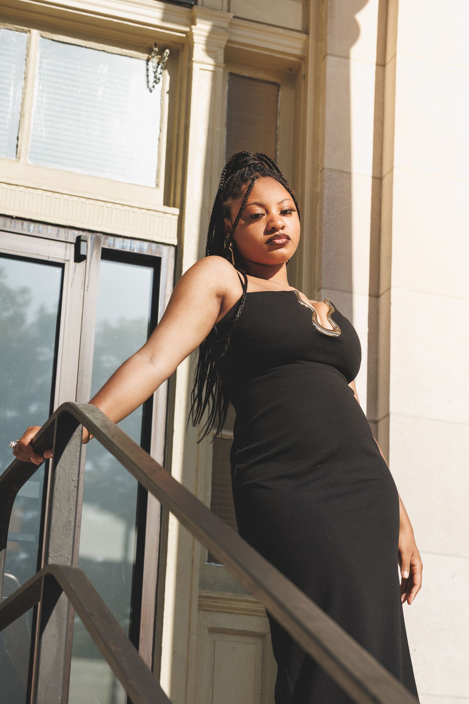 A young Black woman in a black slip dress poses gracefully on sunlit outdoor stairs, leaning on a metal railing.
