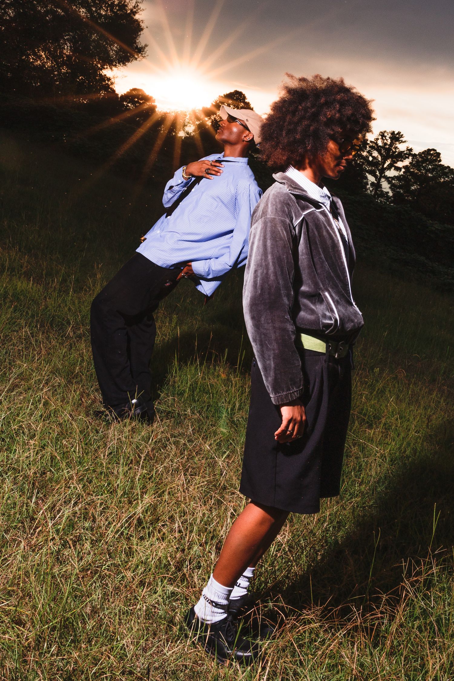 Two stylish individuals pose in a grassy field at golden hour sunset, wearing streetwear fashion.