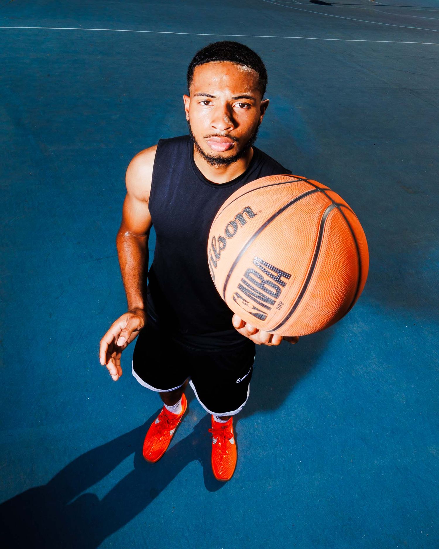 Man holding Wilson NBA basketball on blue outdoor court, wearing black jersey and red sneakers, photographed from above.