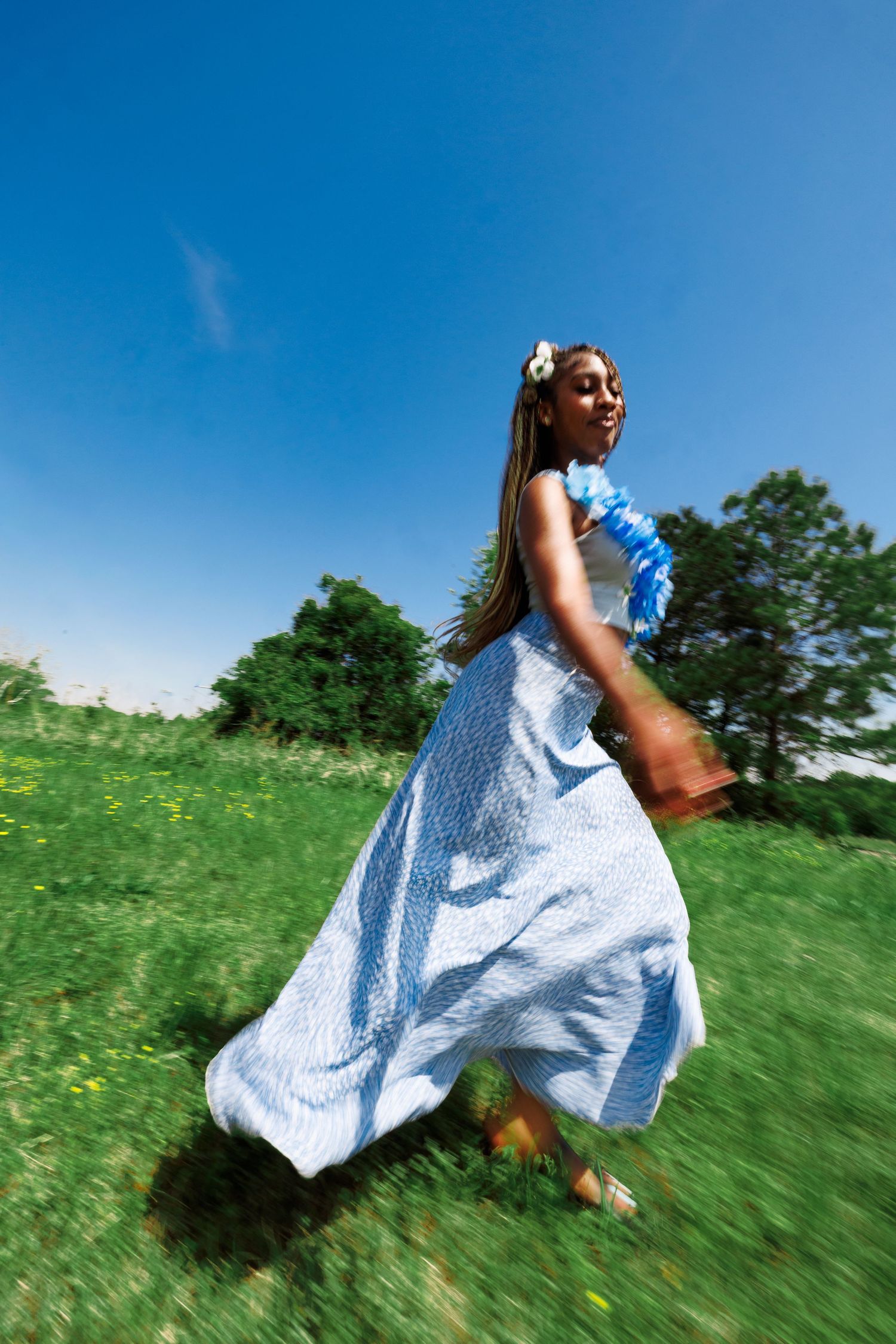 Young woman in flowing blue dress running joyfully through a green meadow under a bright blue sky.