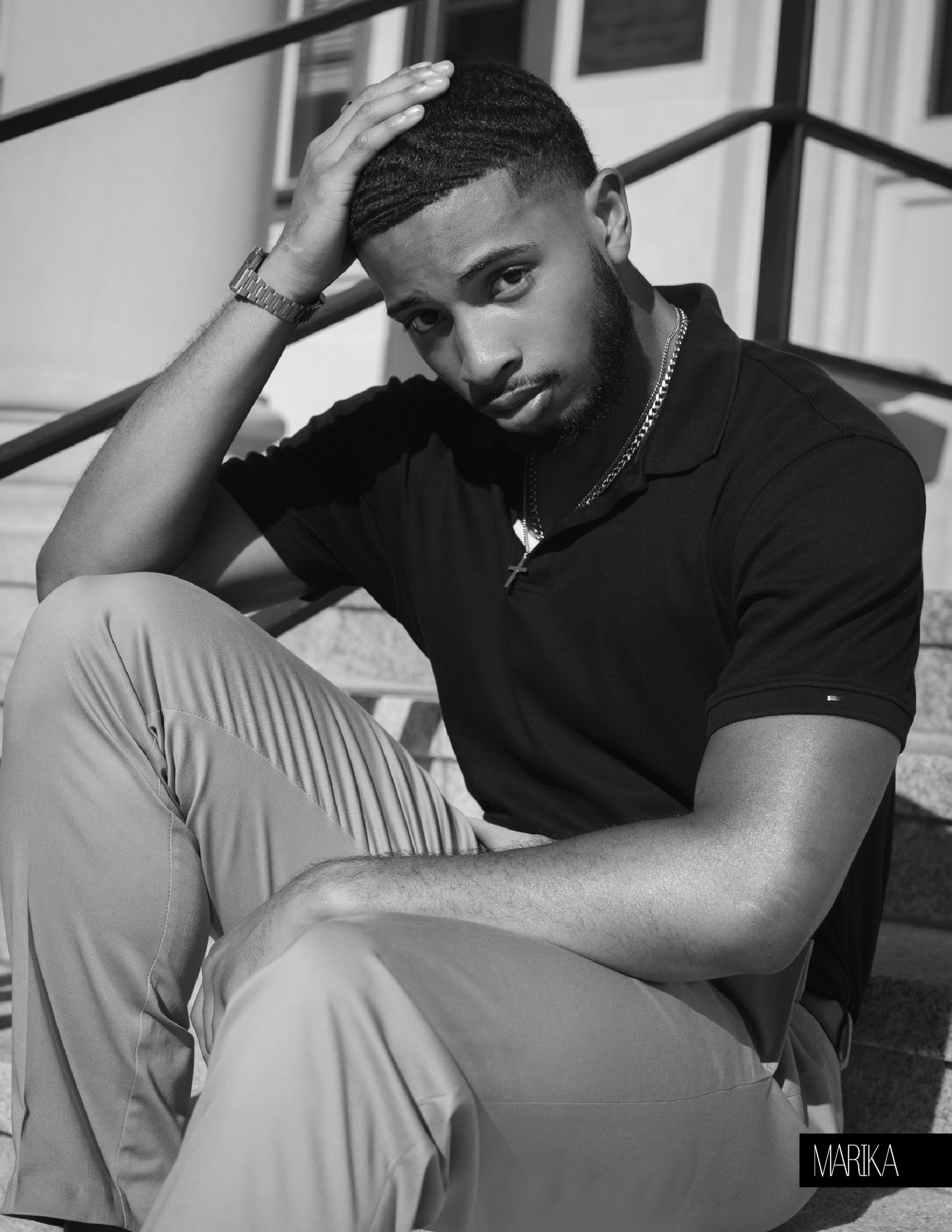 Black and white portrait of a young man in a black polo shirt sitting on steps with hand raised to head.