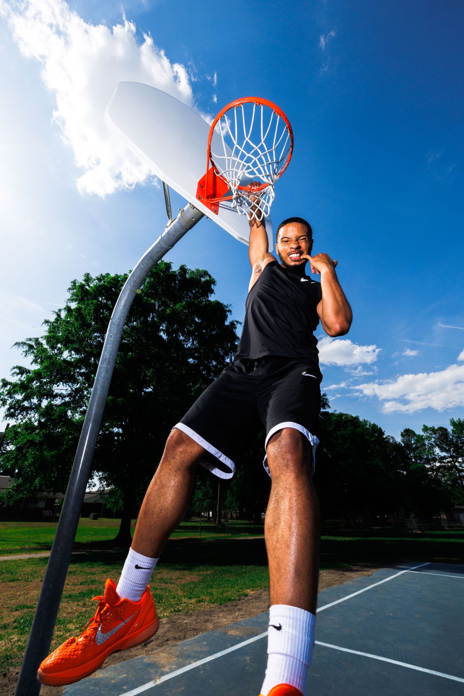 Athletic man dunking basketball on outdoor court, wearing black Nike outfit and orange sneakers under blue sky.