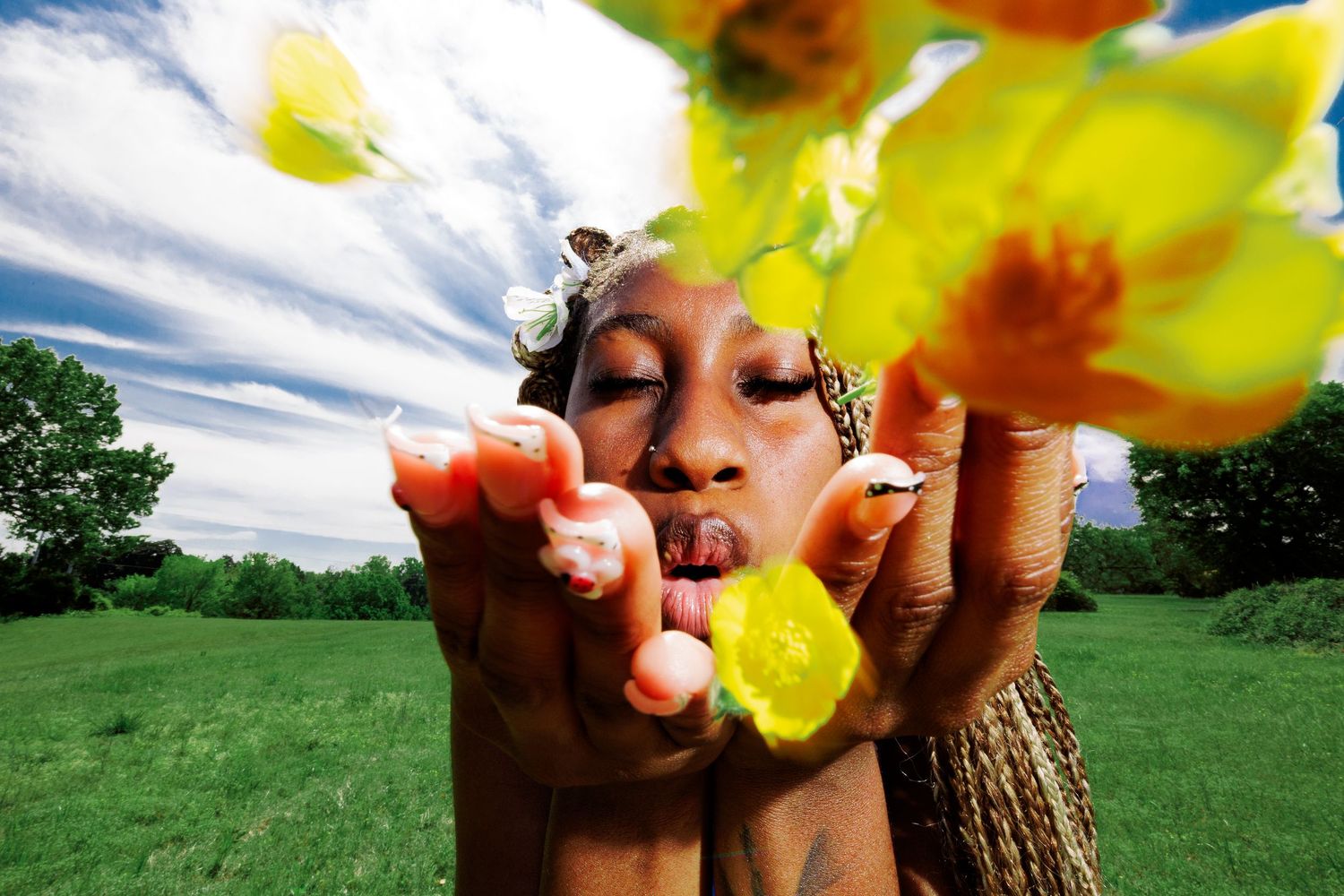 Woman blowing yellow flowers toward camera in a lush green field under a blue sky.