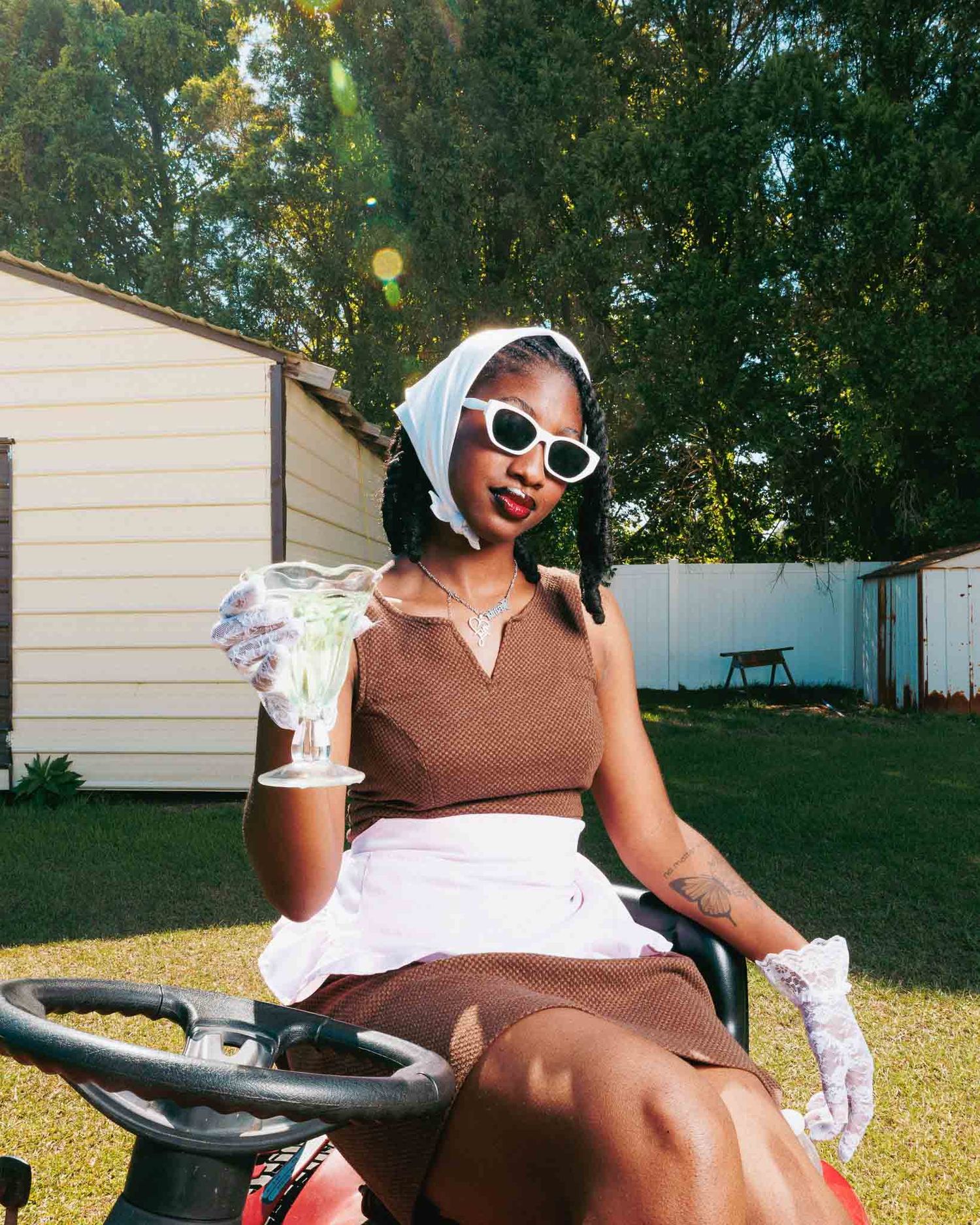 A stylish woman in retro sunglasses and head scarf rides a lawn mower, holding a drink while wearing a brown top and white skirt.