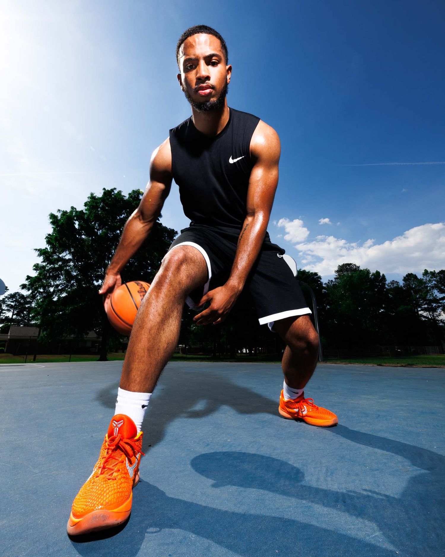A young man in Nike athletic wear poses with a basketball on an outdoor court wearing orange Nike sneakers.